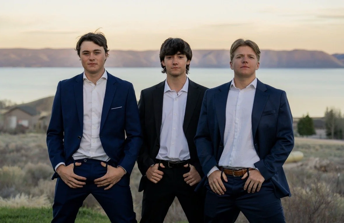 Three young men in suits standing outdoors before a lake and mountains at sunset.