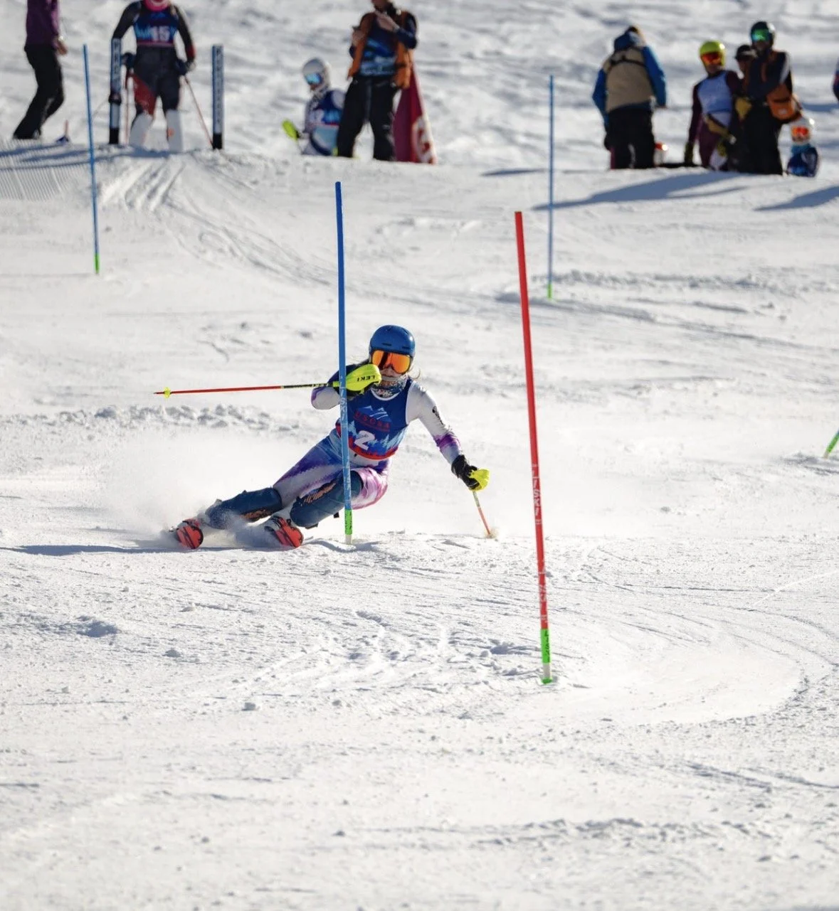 A young skier navigates a slalom course on snow, leaning into a turn while wearing a blue helmet, goggles, and colorful skiing gear, with onlookers in the background.