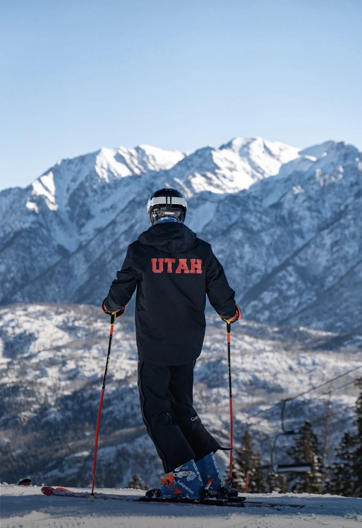 A skier wearing a black jacket with 'UTAH' written on the back, skiing in a snowy mountain landscape with snow-covered peaks and a clear blue sky.