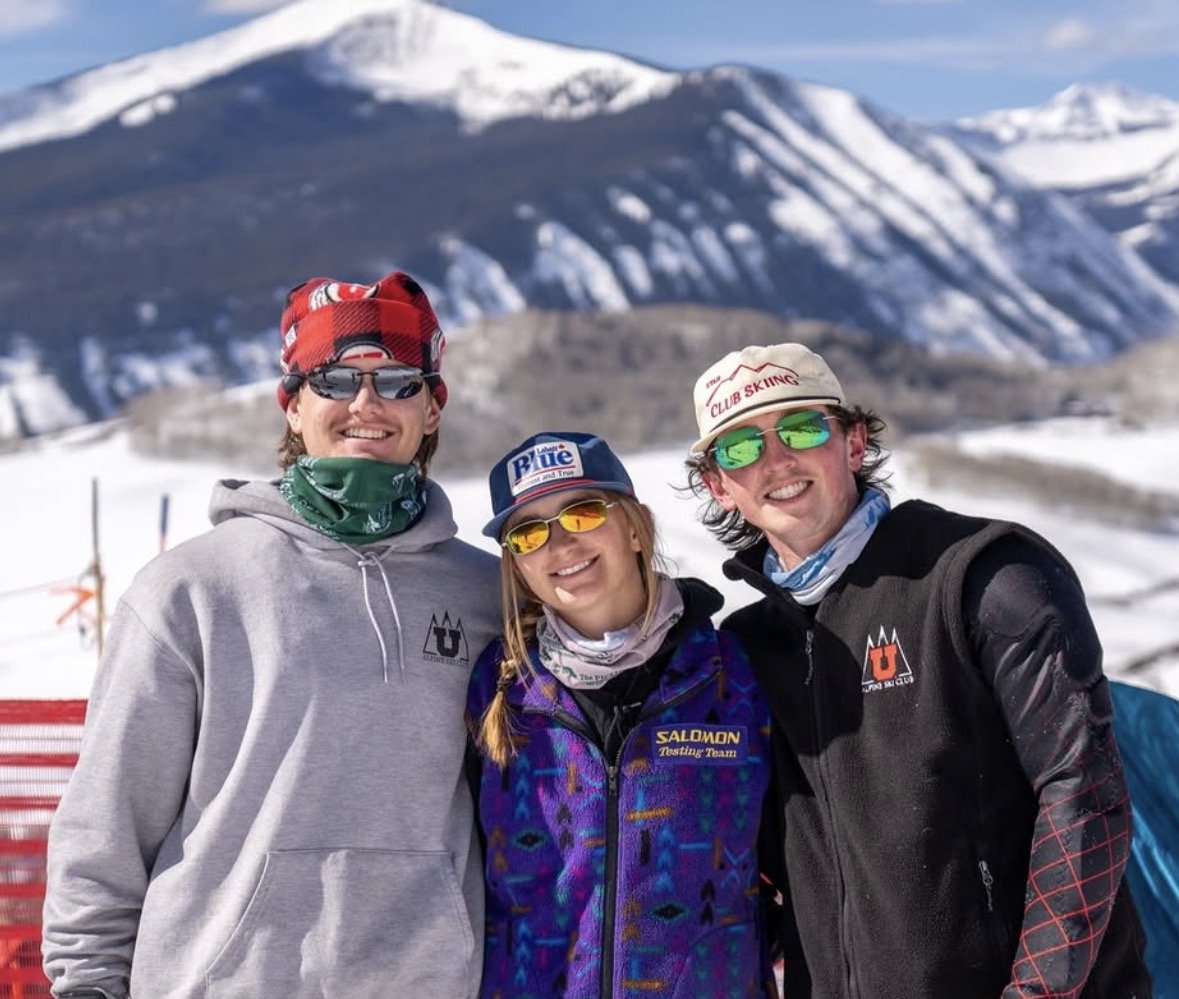 Three people smiling in snowy mountains, wearing sunglasses and winter gear, with mountain peaks in the background.