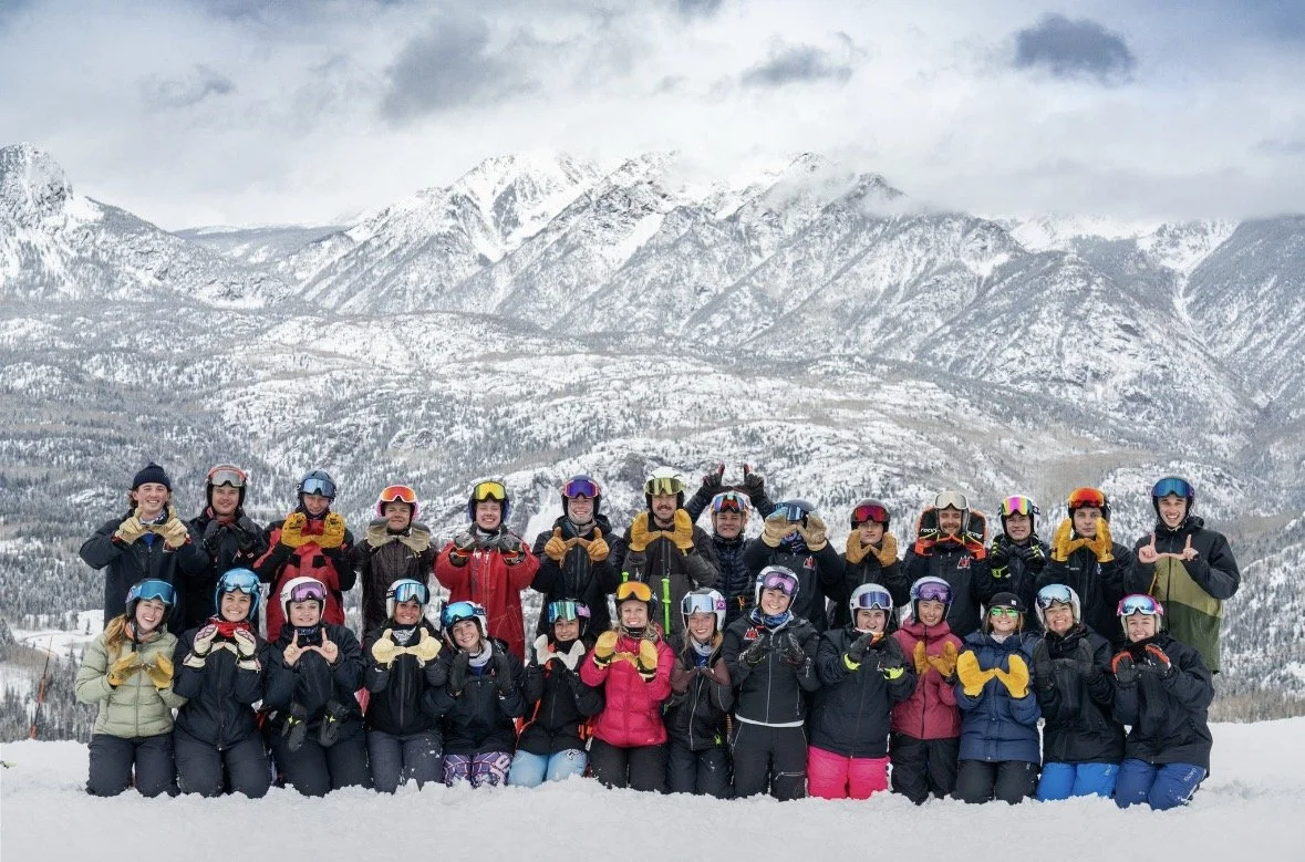 Group of people in snow gear and helmets posing outdoors on a snowy landscape with snow-covered mountains in the background.