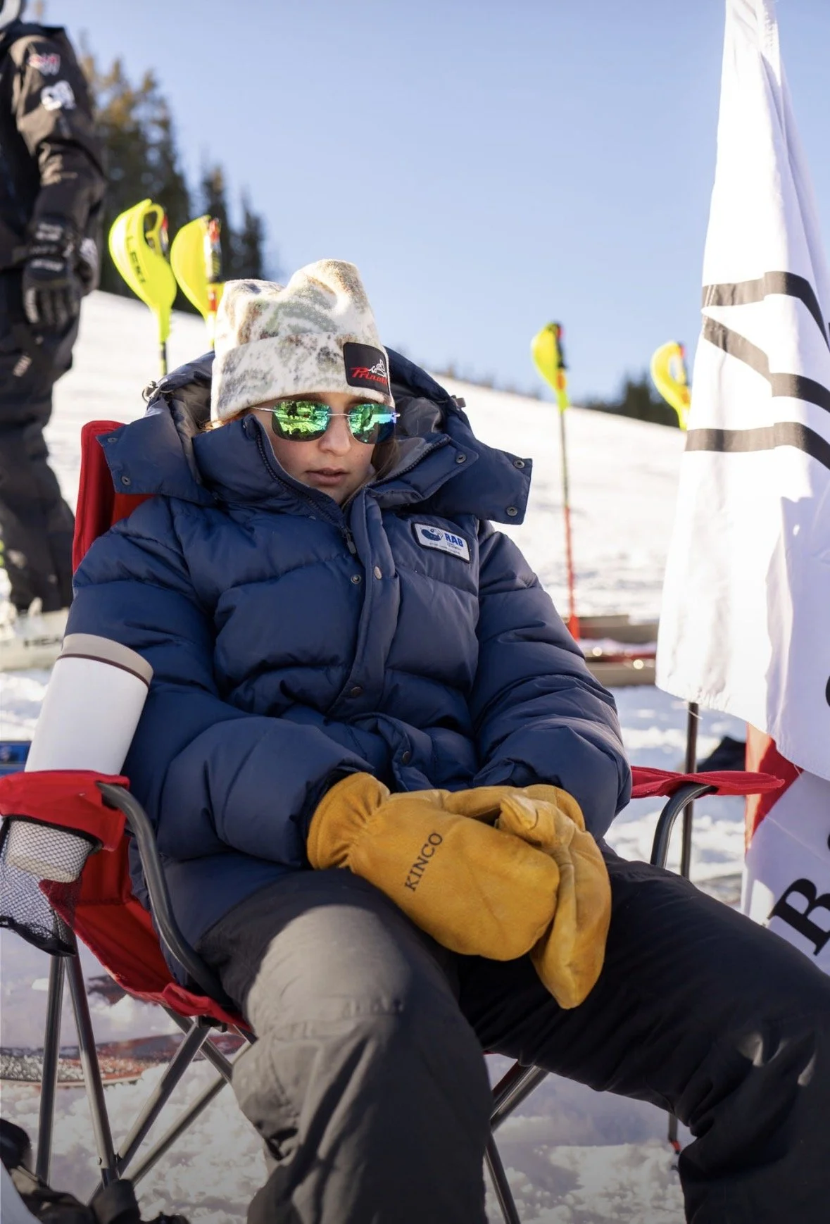 A child wearing a patriotic winter jacket, beanie, reflective sunglasses, and yellow gloves sits outdoors in a snow-covered area, with ski equipment visible in the background.