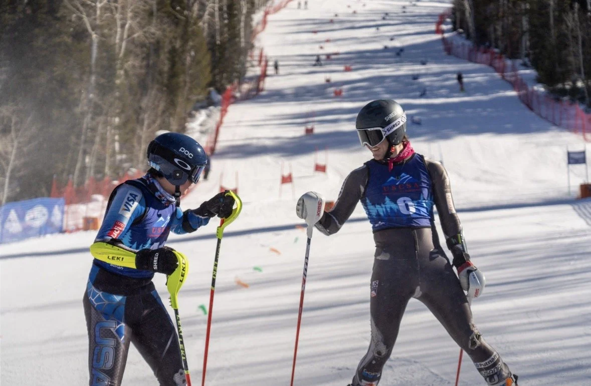 Two young skiers talking on a snow-covered ski slope during a race, with trees and a red safety fence in the background.
