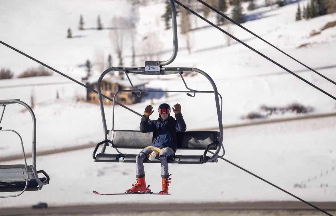 A person wearing ski gear and red ski boots on a ski lift, raising their hands while sitting on a ski lift chair in a snowy mountain landscape.