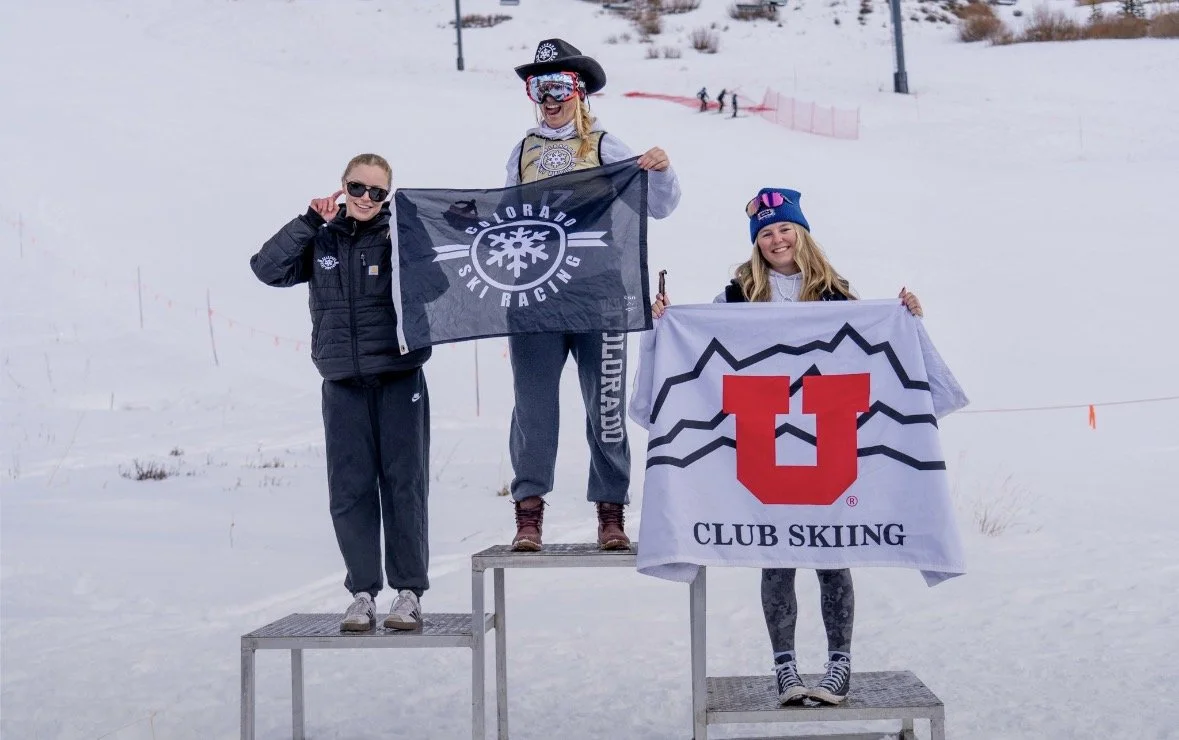 Three women standing on a snowy stage holding flags, celebrating at a ski race event.