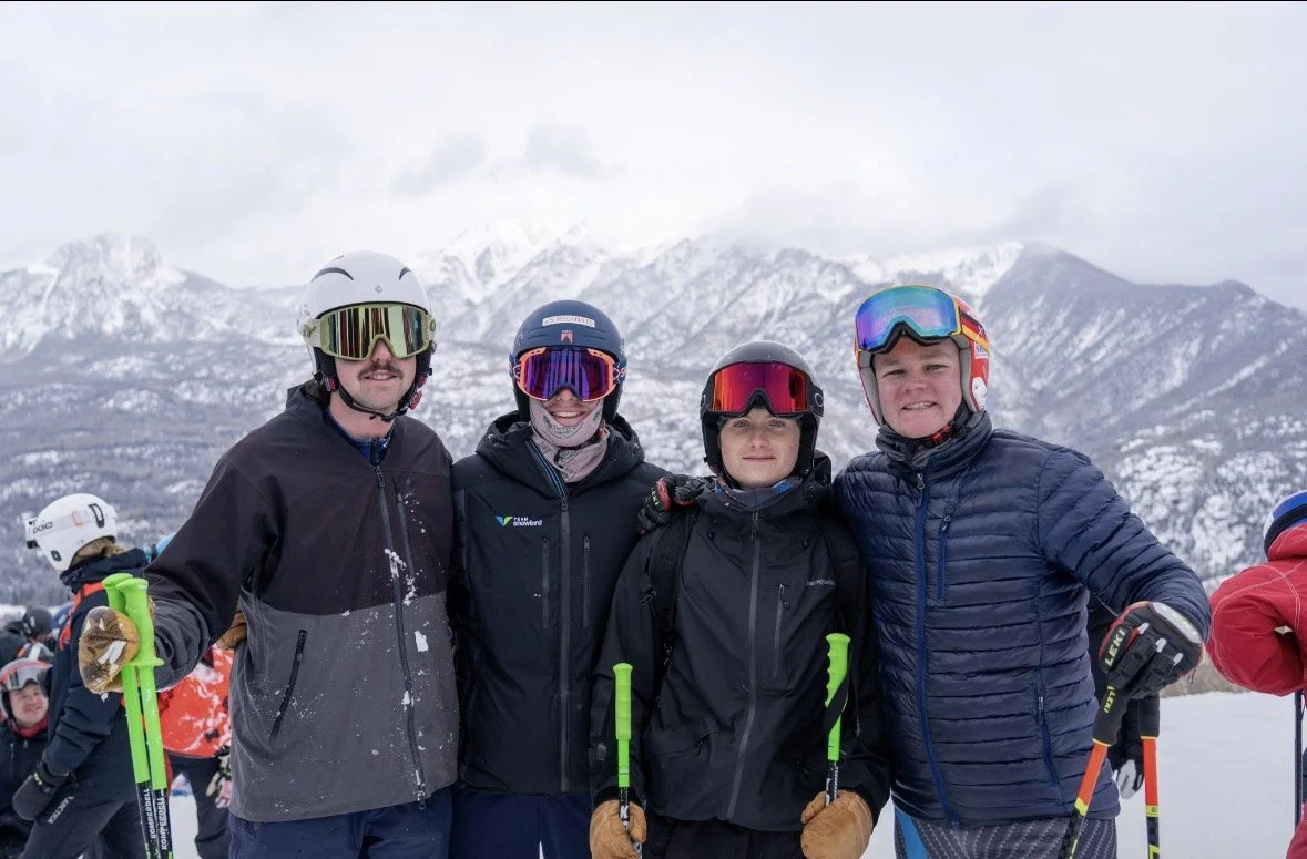 Four people wearing ski gear and helmets standing together in a snowy mountain landscape.