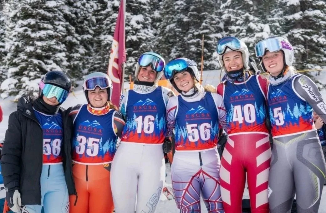 Six female athletes in ski uniforms and helmets, standing together outdoors in a snowy forest, smiling after a winter sports event.