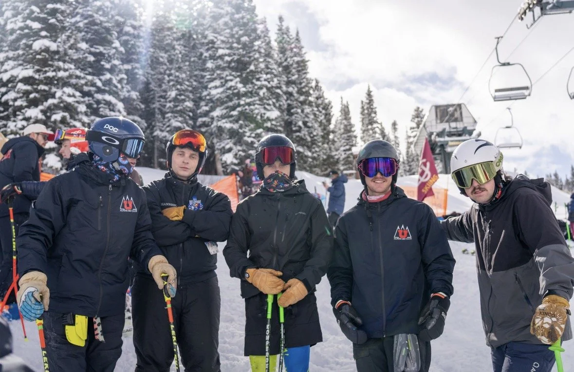 Group of five men dressed in skiing gear, standing outdoors in a snowy ski resort with person skiing chairs and snow-covered trees in the background.