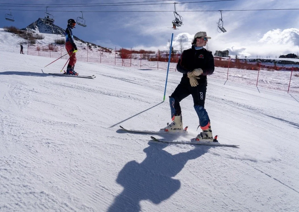 A skier standing on snowy slope with ski poles, wearing black jacket, helmet, and goggles, underneath ski lift with empty chairs, in a ski resort with buildings and mountains in the background.