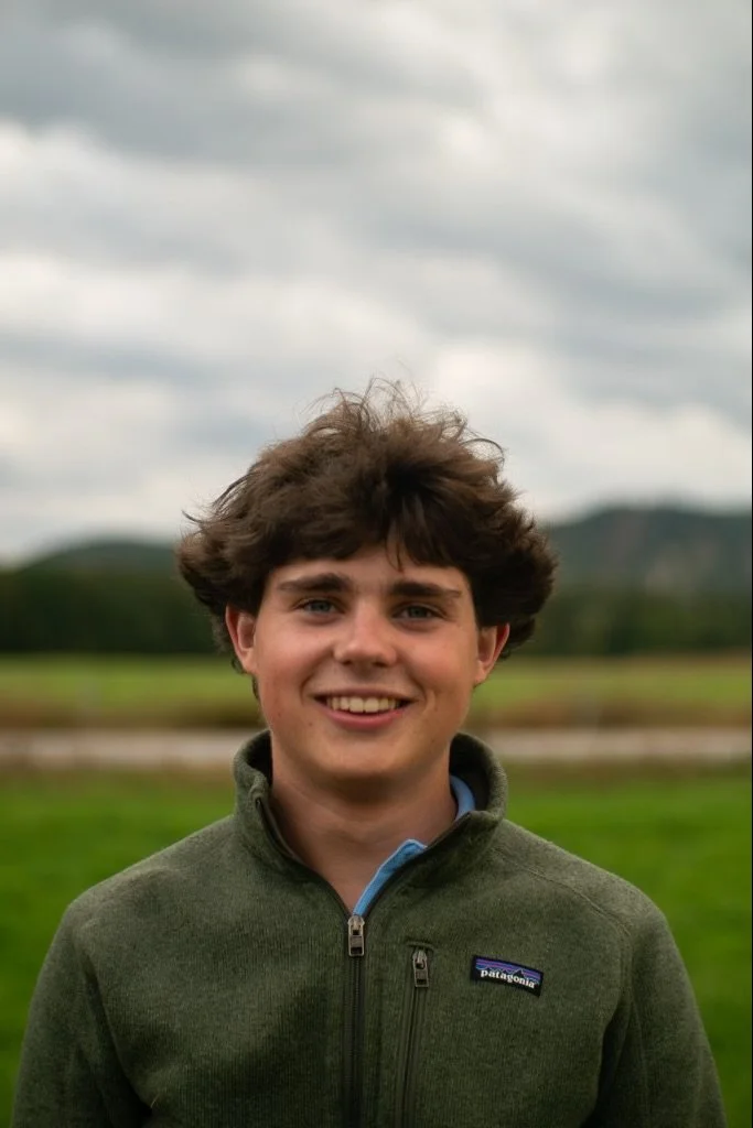 A young man with curly brown hair smiling outdoors, wearing a green Patagonia jacket, with a blurred green landscape and cloudy sky in the background.