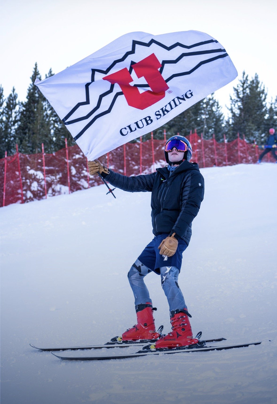 A person skiing on snow while holding a flag with the logo of Club Skiing. The skier is wearing a black jacket, patterned ski pants, red ski boots, and a helmet with goggles. There are trees and a red safety fence in the background.