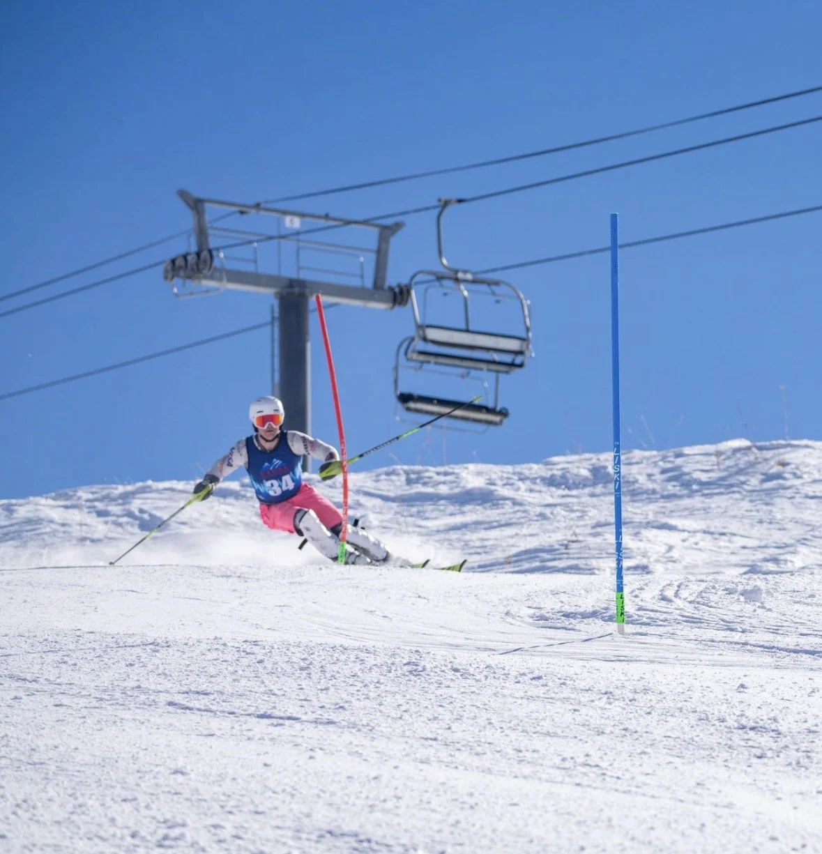 A skier in a pink skirt and white top, wearing a helmet and goggles, skiing down a snow-covered slope under a chairlift on a sunny day.