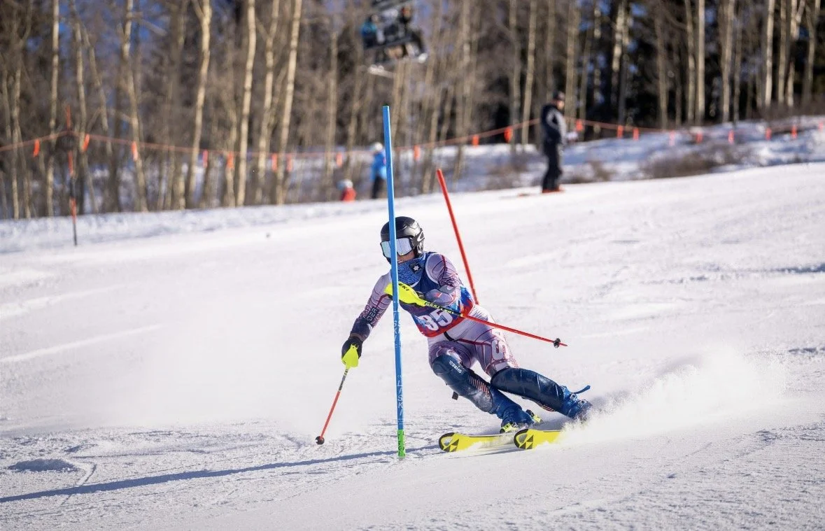 A skier competes in a downhill race, navigating around a blue slalom gate on a snow-covered slope with trees in the background.