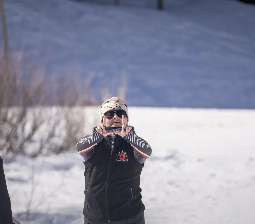 A person wearing sunglasses, a beanie, and a black jacket making a framed camera gesture with their hands outdoors in a snowy landscape.
