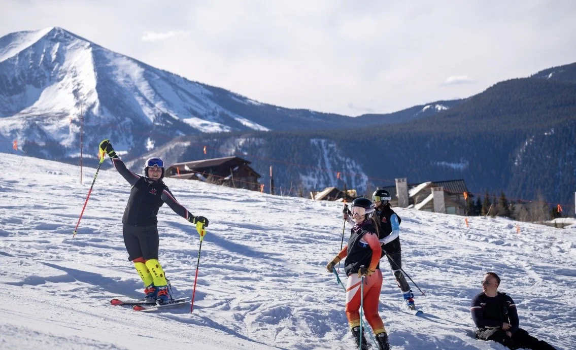 Four skiers on a snowy mountain slope, with mountains and ski lodges in the background, enjoying a sunny day.