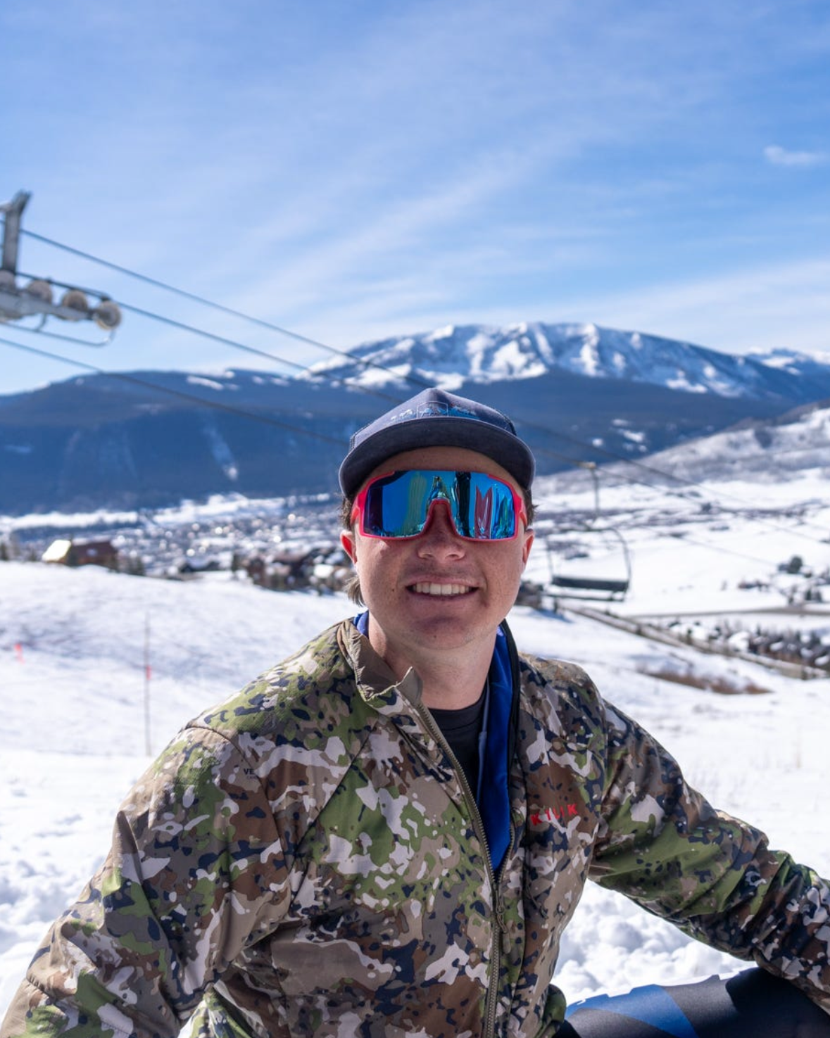 A man wearing a camouflage jacket, a black cap, and ski goggles smiling in a snowy mountain landscape with ski lifts and a mountain in the background.