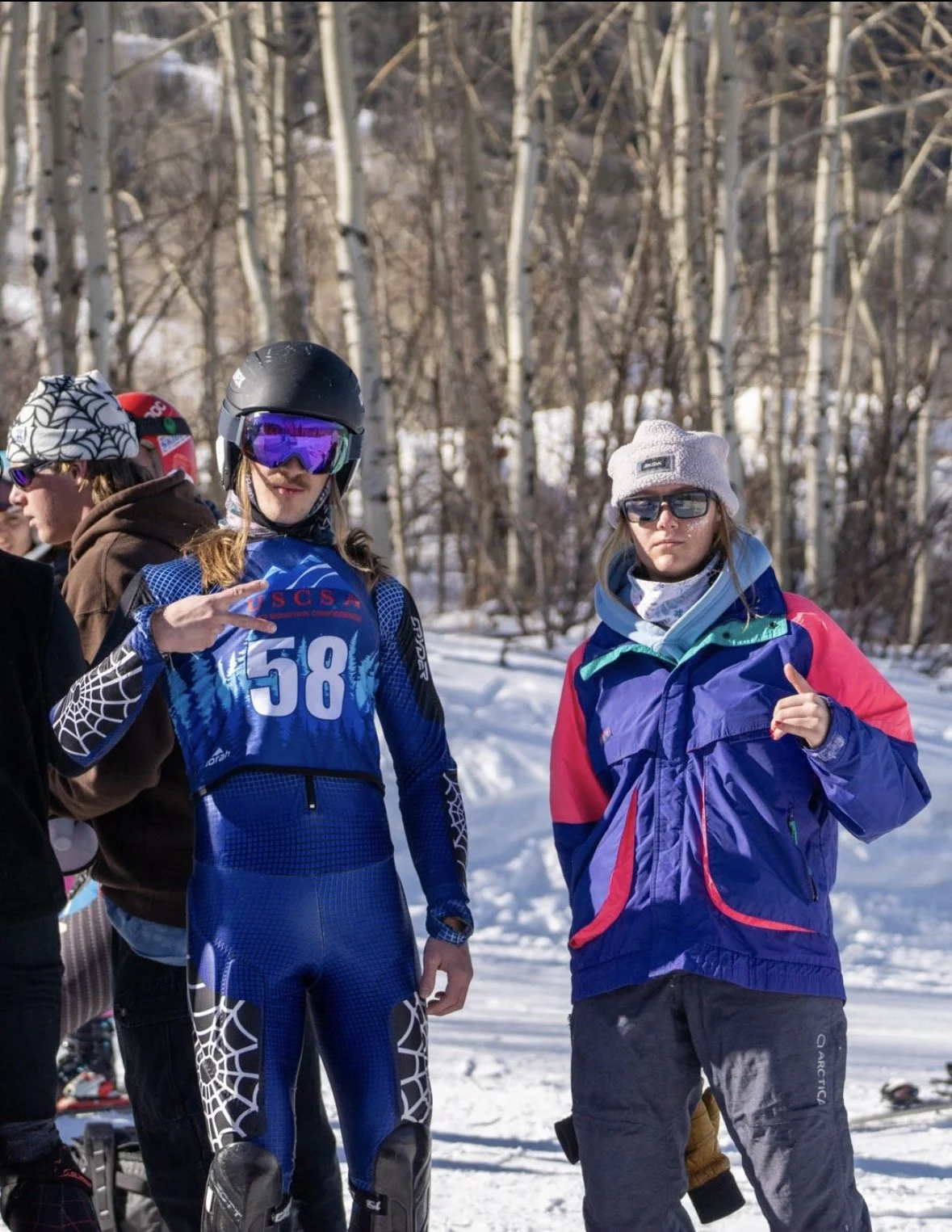 Two young women in winter gear standing outdoors on snow, with a group of people and leafless trees in the background; one is wearing blue sportswear with a helmet and goggles, making a peace sign, and the other is wearing a colorful jacket, beanie, 
