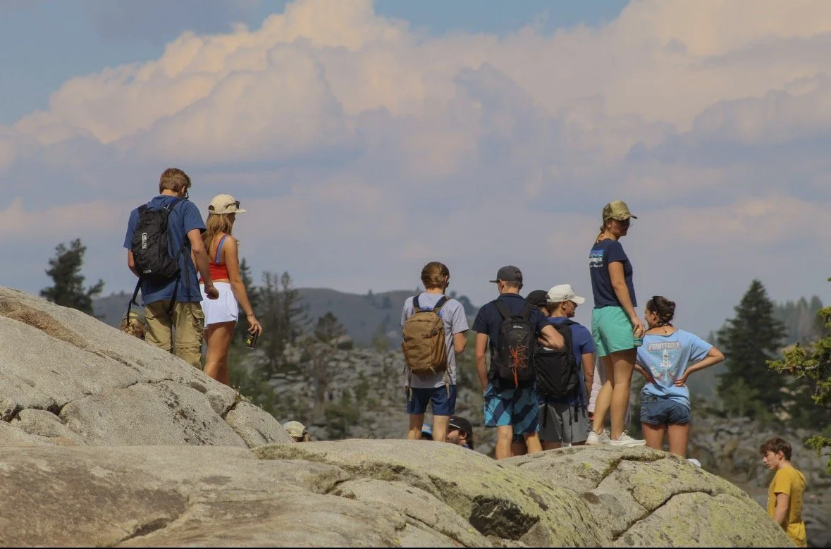 Group of children and teenagers hiking on a rocky terrain in a mountainous area with trees and a cloudy sky in the background.