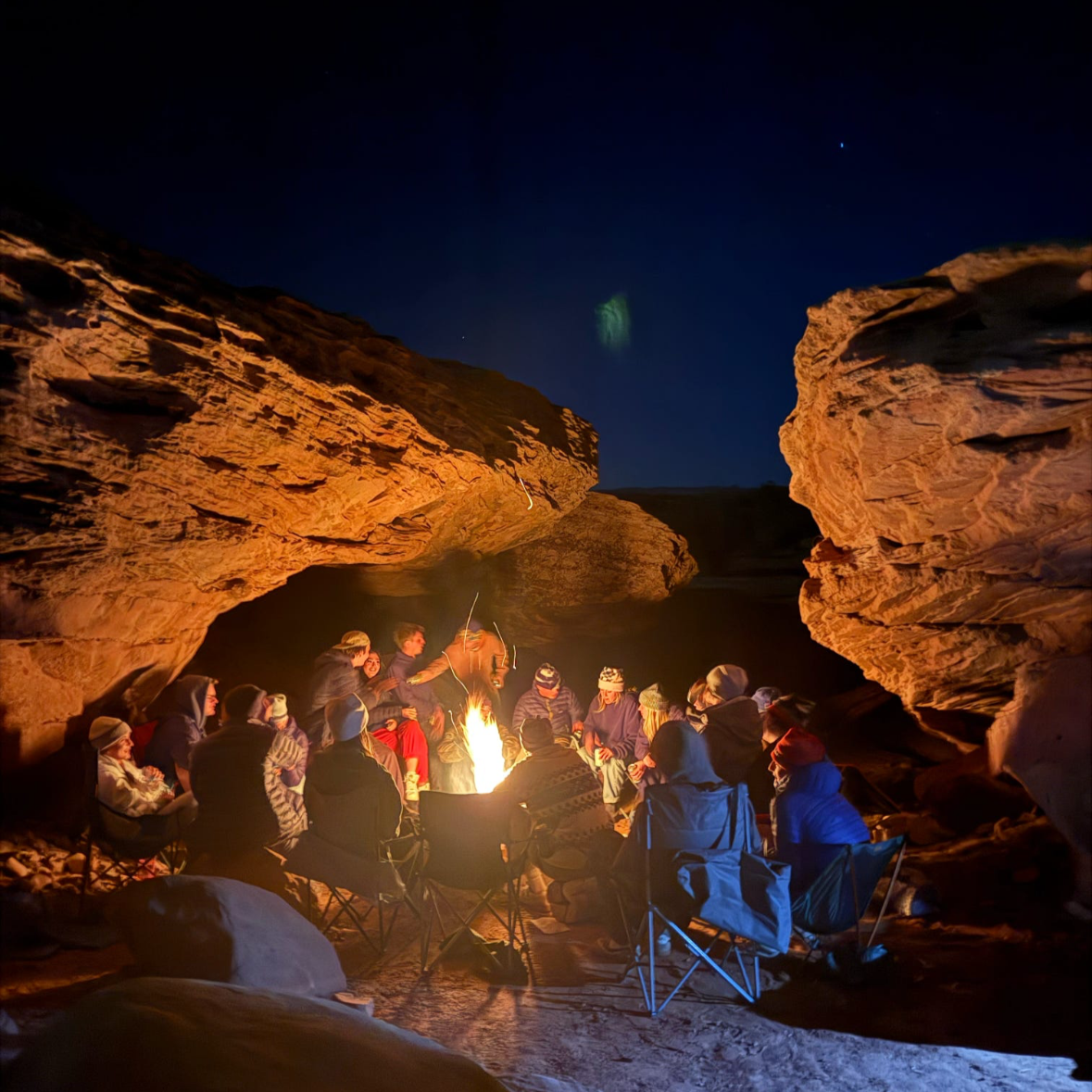 Group of people gathered around a campfire in a rocky outdoor canyon at night, with a clear starry sky above.