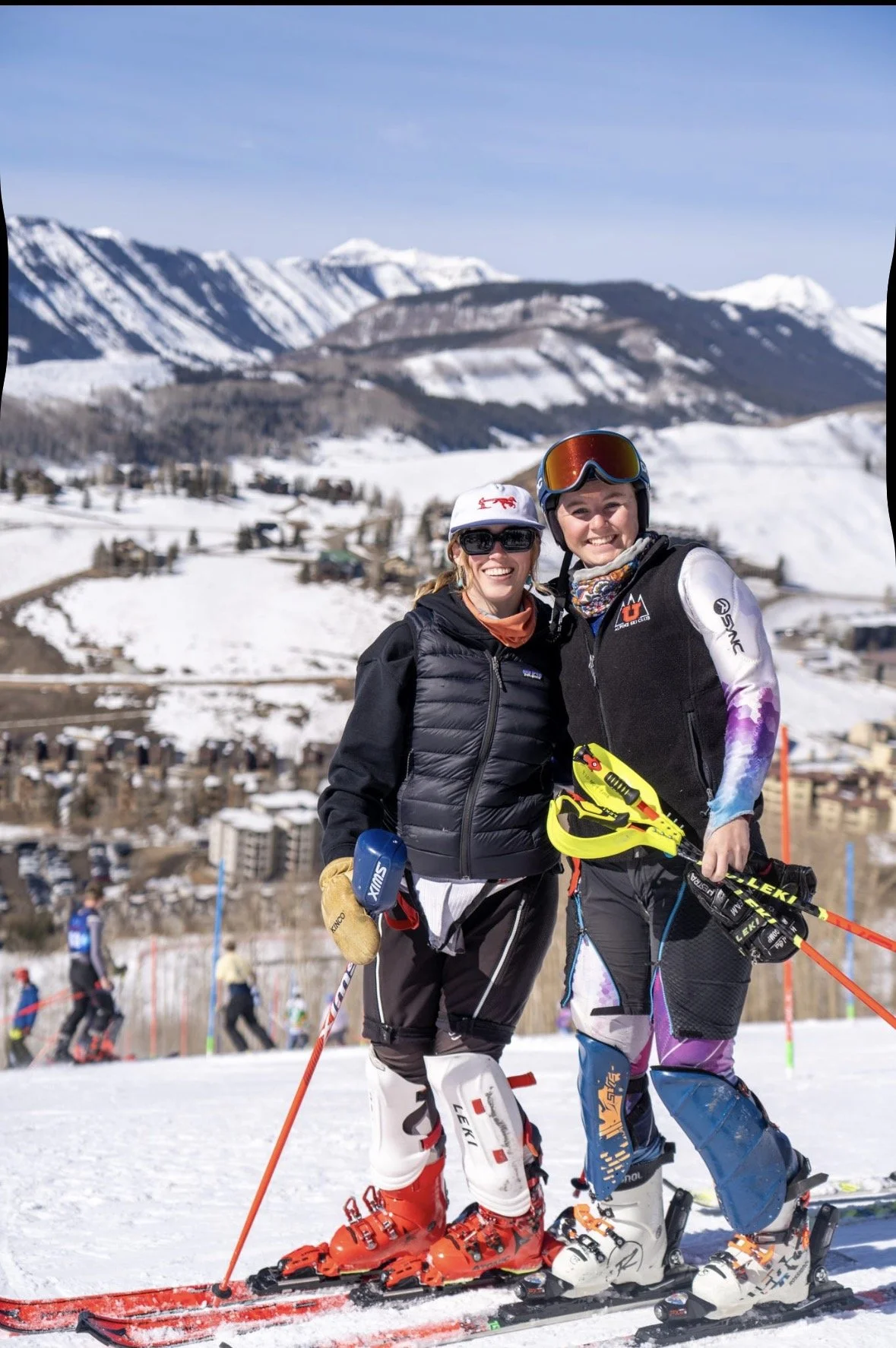 Two women smiling on skis in a snowy mountain landscape with a town below and snow-capped mountains in the background.