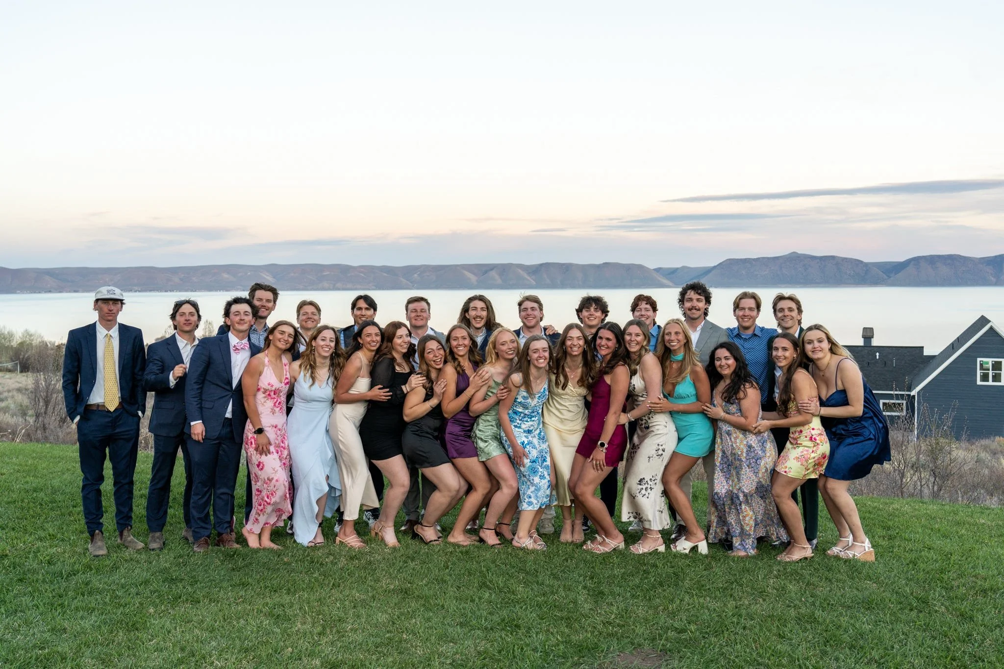 A large group of young adults dressed in formal attire standing outdoors on a grassy area near water with mountains in the background, smiling and posing for a group photo.