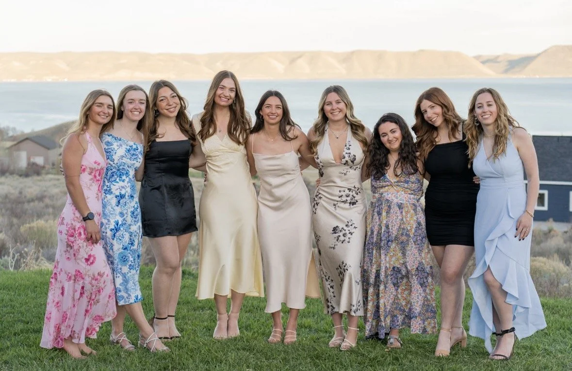 Group of ten women standing outdoors on grass, dressed in colorful and elegant dresses, with a backdrop of water and distant hills.