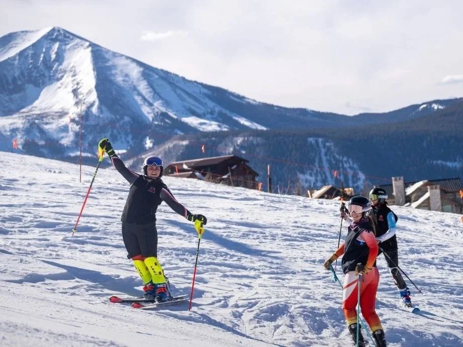 Three people skiing on a snowy mountain with a mountain range in the background.