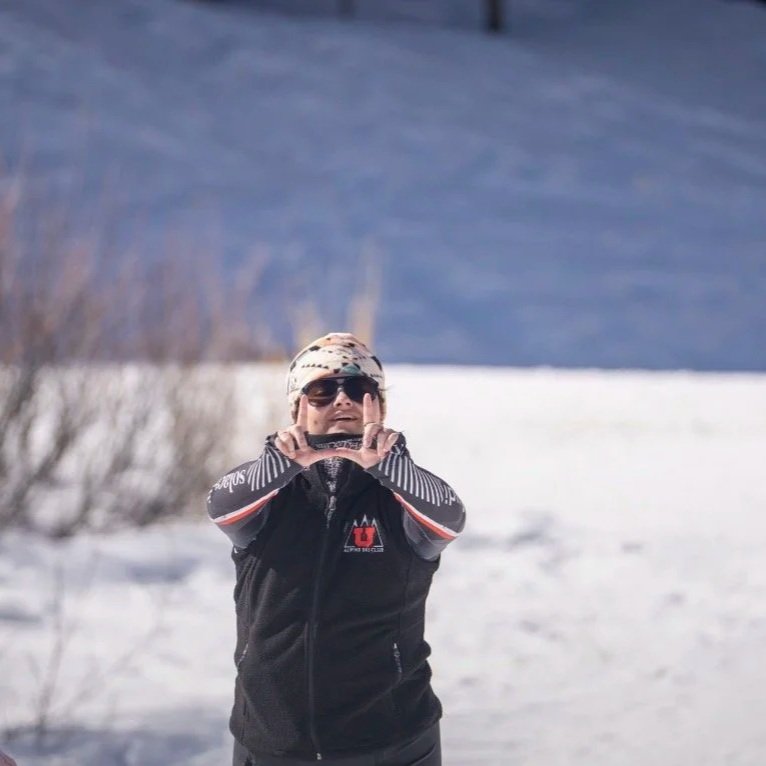 Person wearing sunglasses, a patterned hat, and a black jacket with red and white accents, standing outdoors in a snowy landscape.