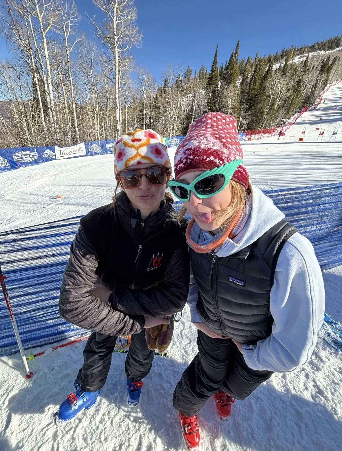 Two women standing on a snowy ski slope, wearing sunglasses, beanies, and winter jackets, posing for a photo amidst a mountain landscape with trees, ski run, and blue sky.