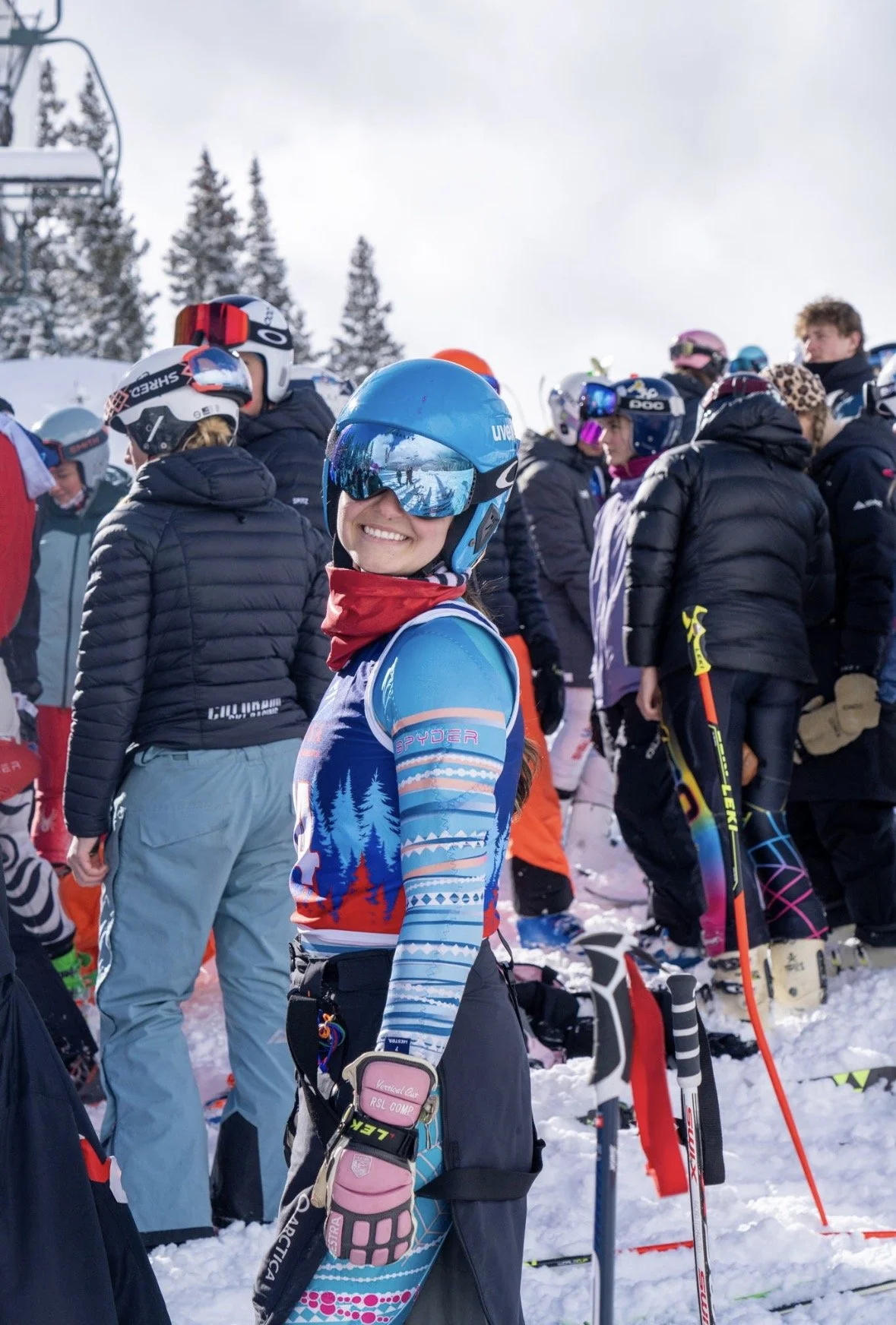 Smiling female skier in blue and patterned ski suit with goggles, surrounded by other skiers in winter gear at a snowy mountain resort.