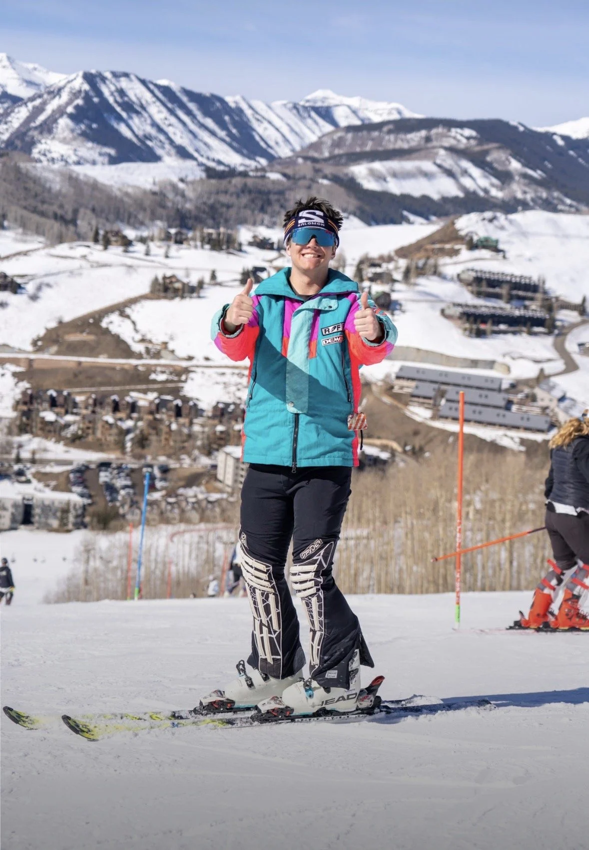A person in colorful winter sports gear smiling and giving a thumbs-up on a ski slope with snow-covered mountains in the background.
