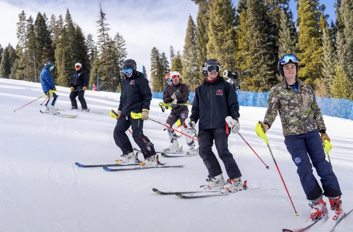 Group of six skiers standing on a snowy slope with trees in the background, some adjusting their ski gear, wearing helmets and winter clothing.