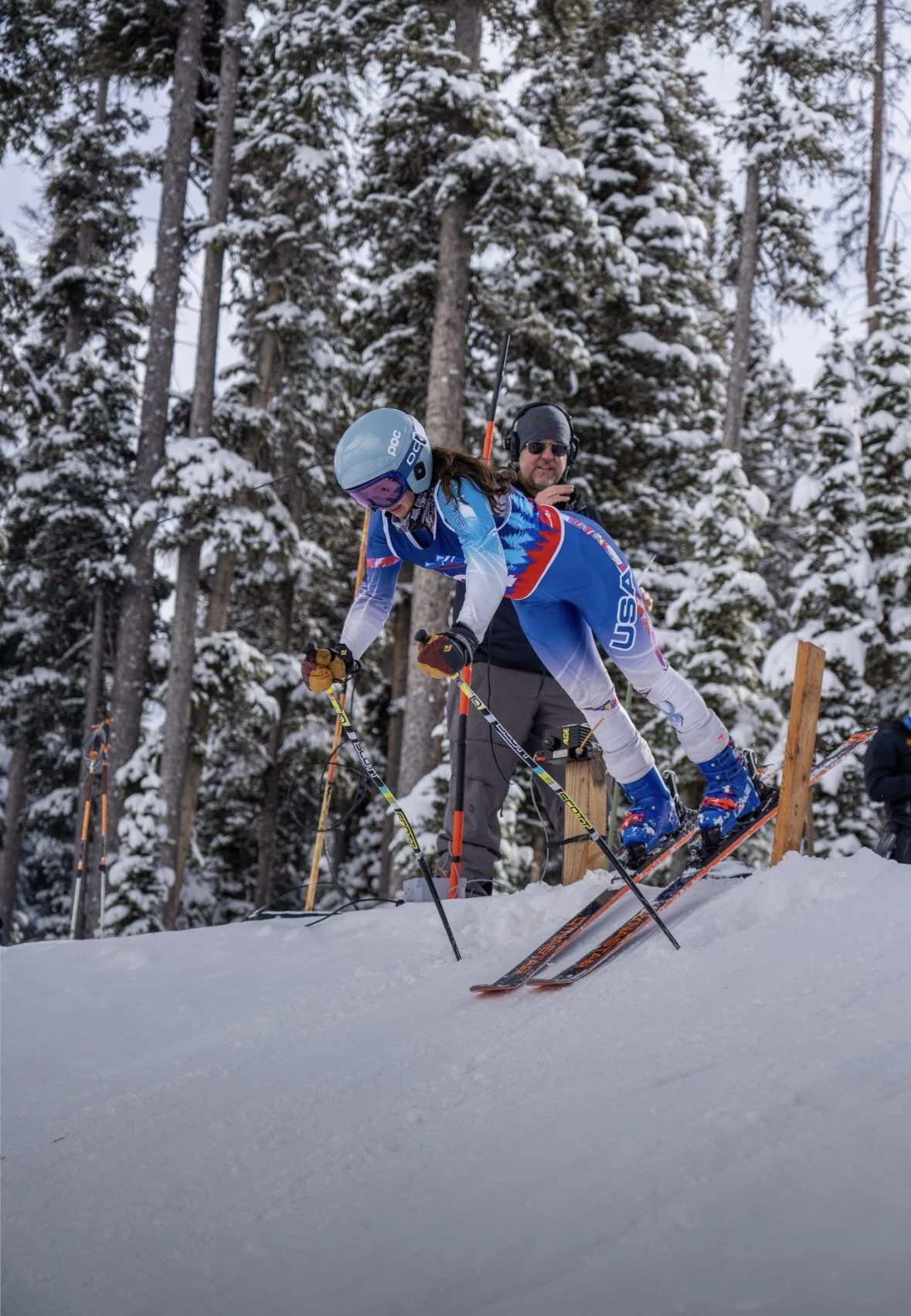 A skier in a blue and white USA team uniform is getting ready to go downhill, with a person in black standing behind in a snowy forested area.