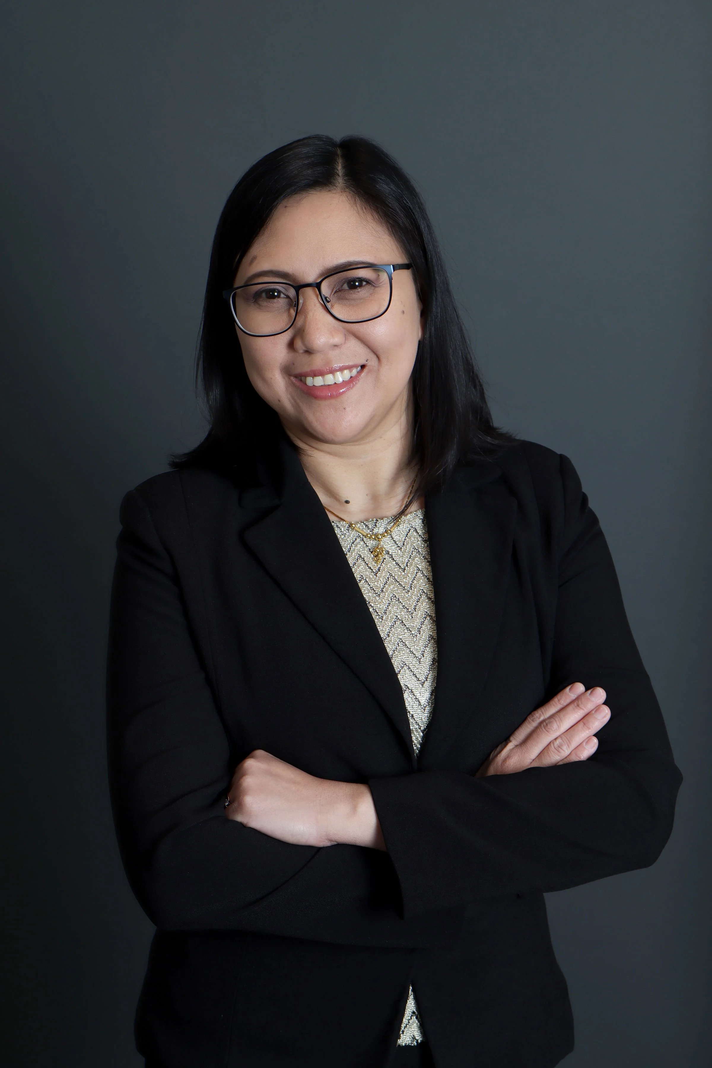 A woman with black hair, wearing glasses and a black blazer, smiling with arms crossed, against a dark gray background.