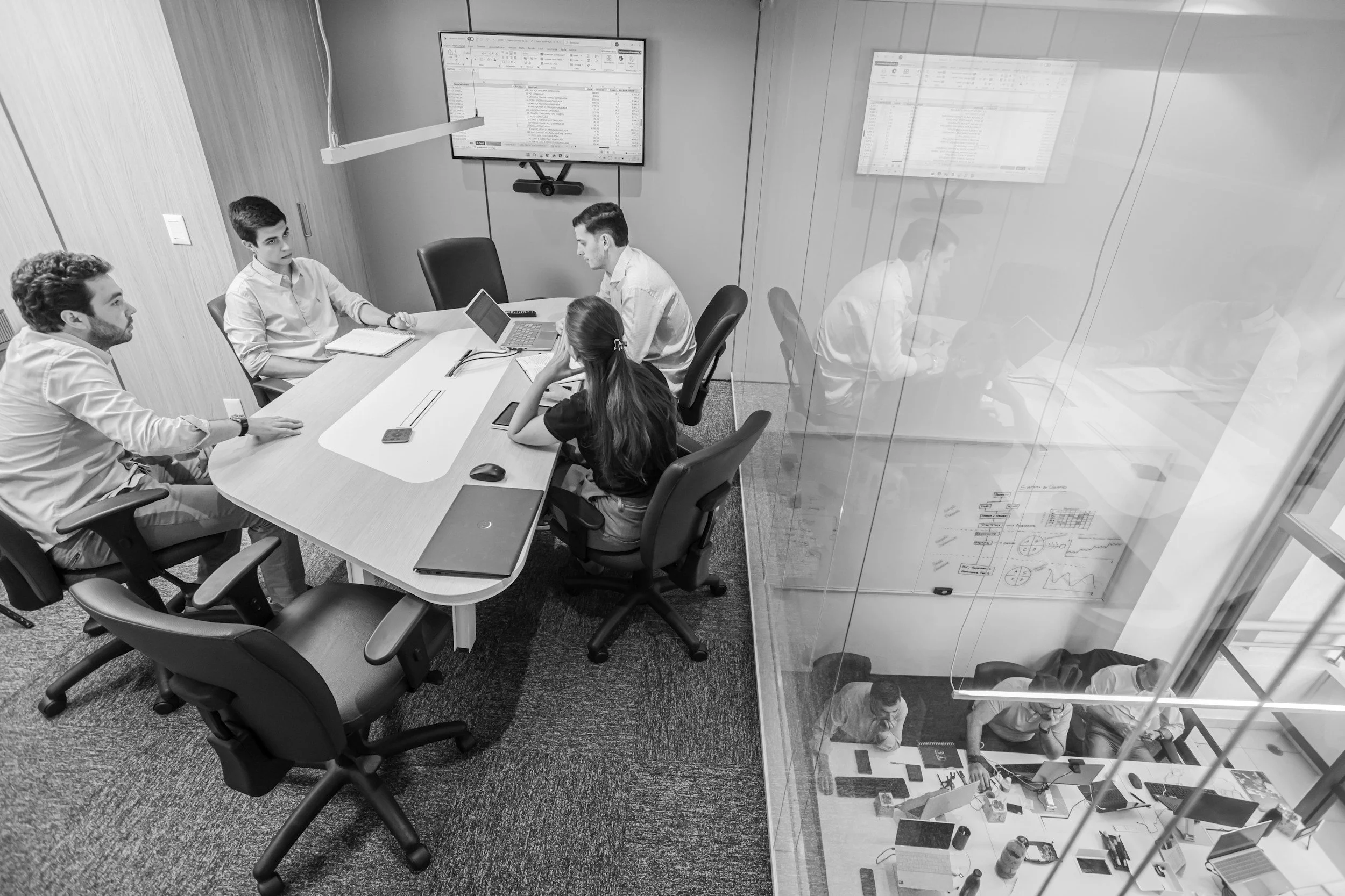 A black and white photo of a modern conference room with five people seated around a rectangular table engaged in a meeting, and two large screens on the wall displaying spreadsheets or data.