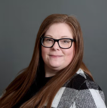 A woman with long red hair wearing glasses and a black-and-white patterned sweater against a gray background.