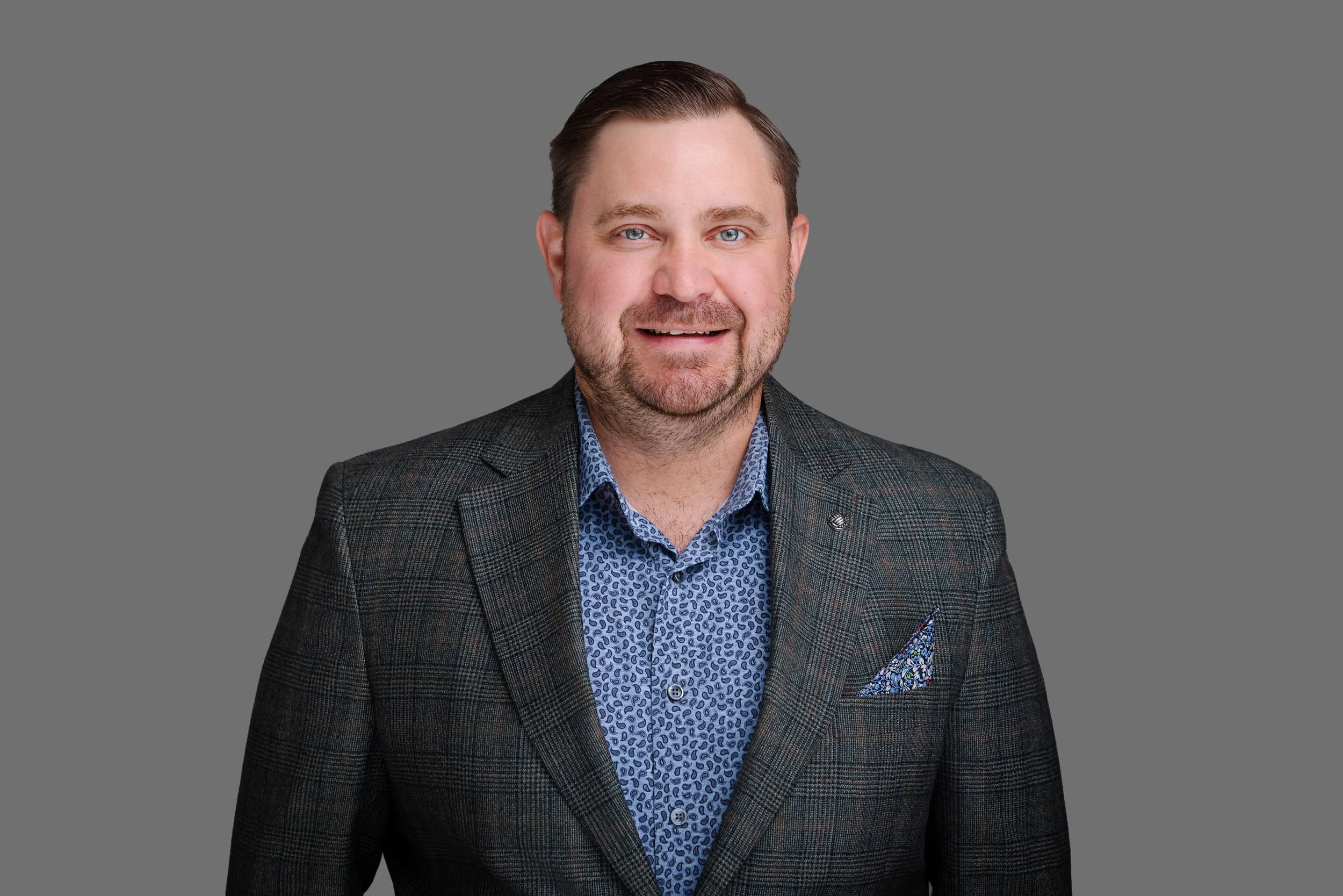 A man with short brown hair and a beard, wearing a navy blue shirt with a white geometric pattern, smiling and posing against a gray background.