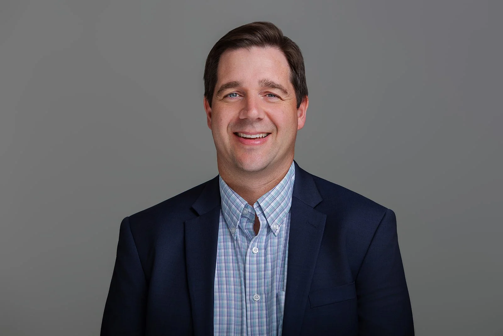 Professional headshot of a smiling man with short dark hair, wearing a suit jacket and checkered shirt, against a gray background.
