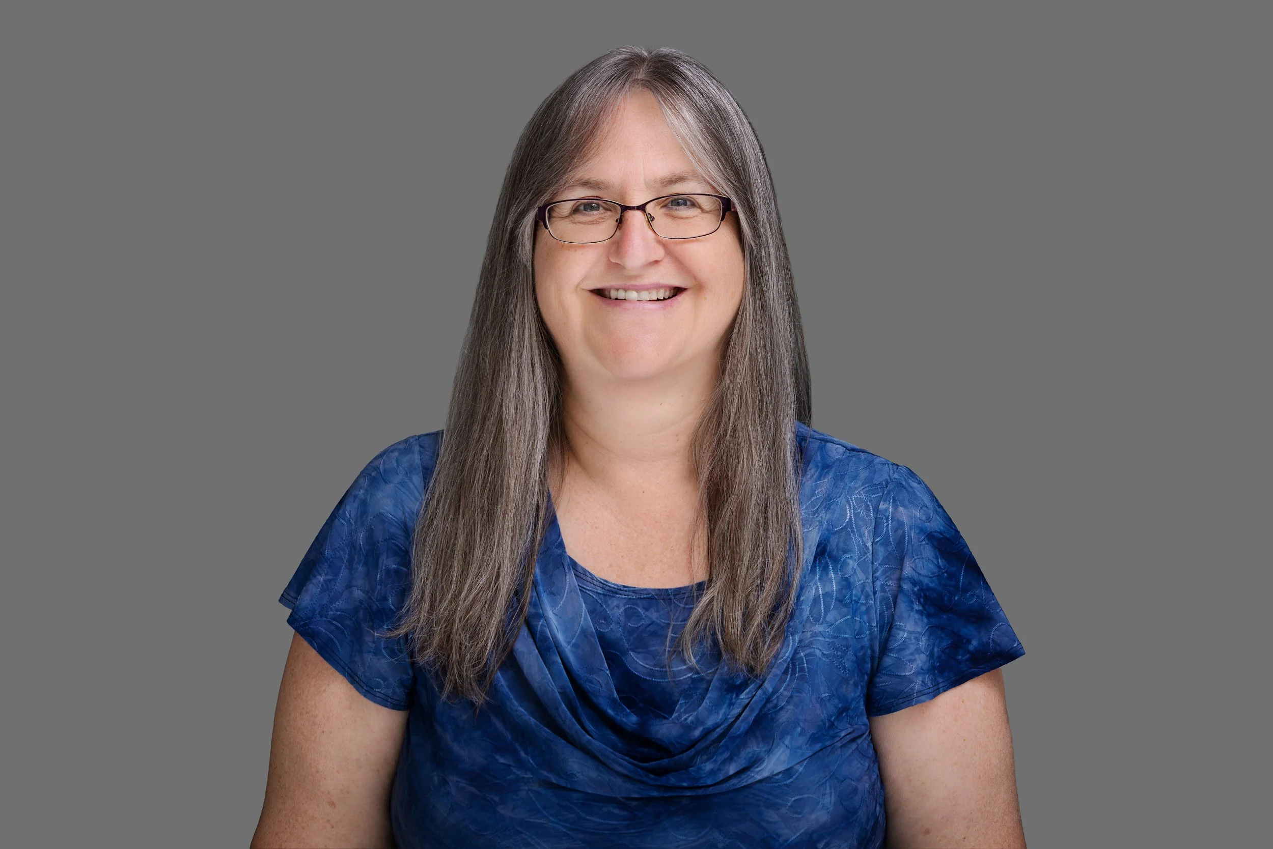 Portrait of a woman with long gray hair, glasses, and a blue patterned blouse, smiling against a gray background.