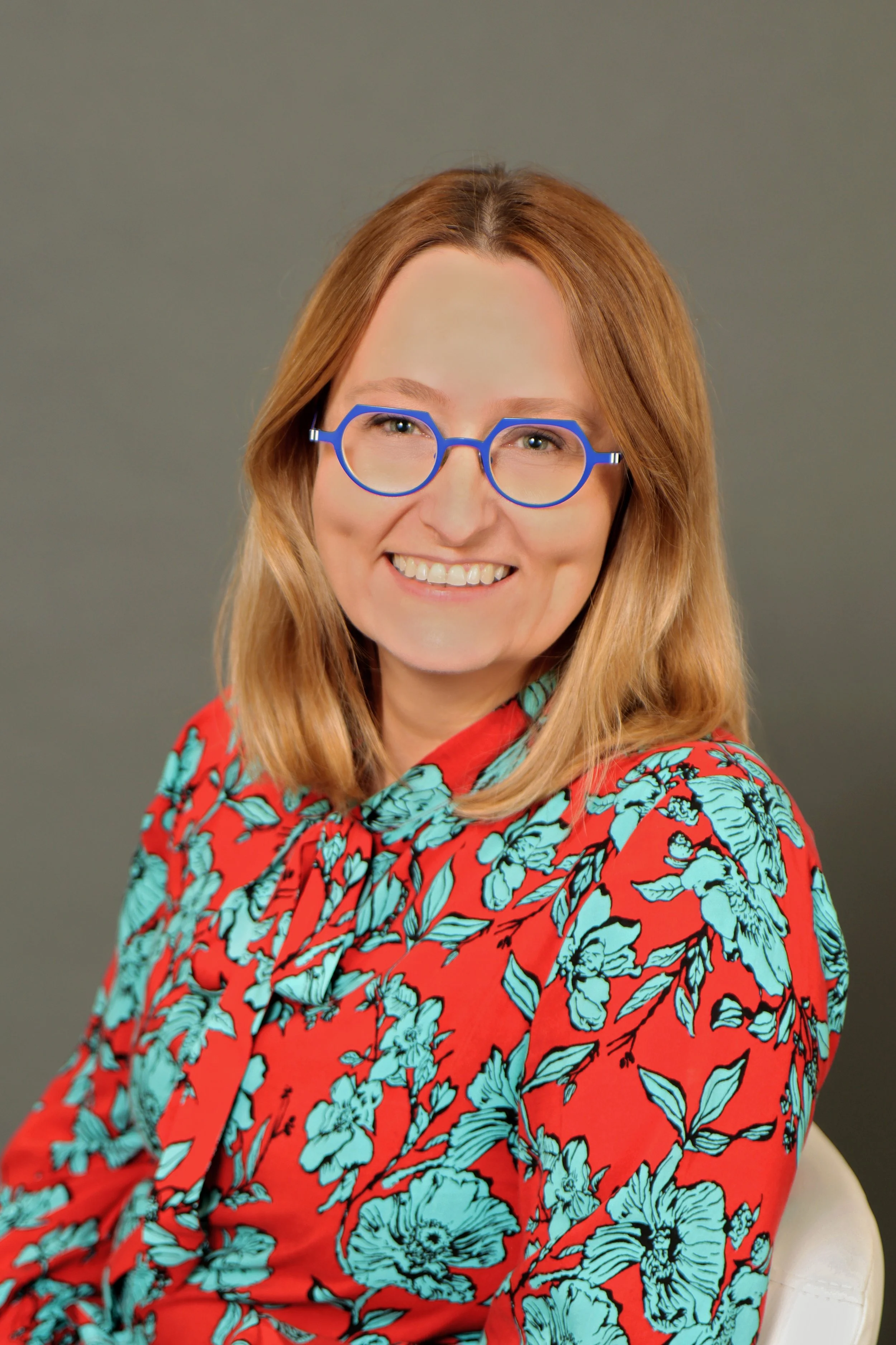A woman with shoulder-length red hair and glasses with blue frames, smiling and wearing a red dress with a blue floral pattern, sitting on a white chair against a plain gray background.