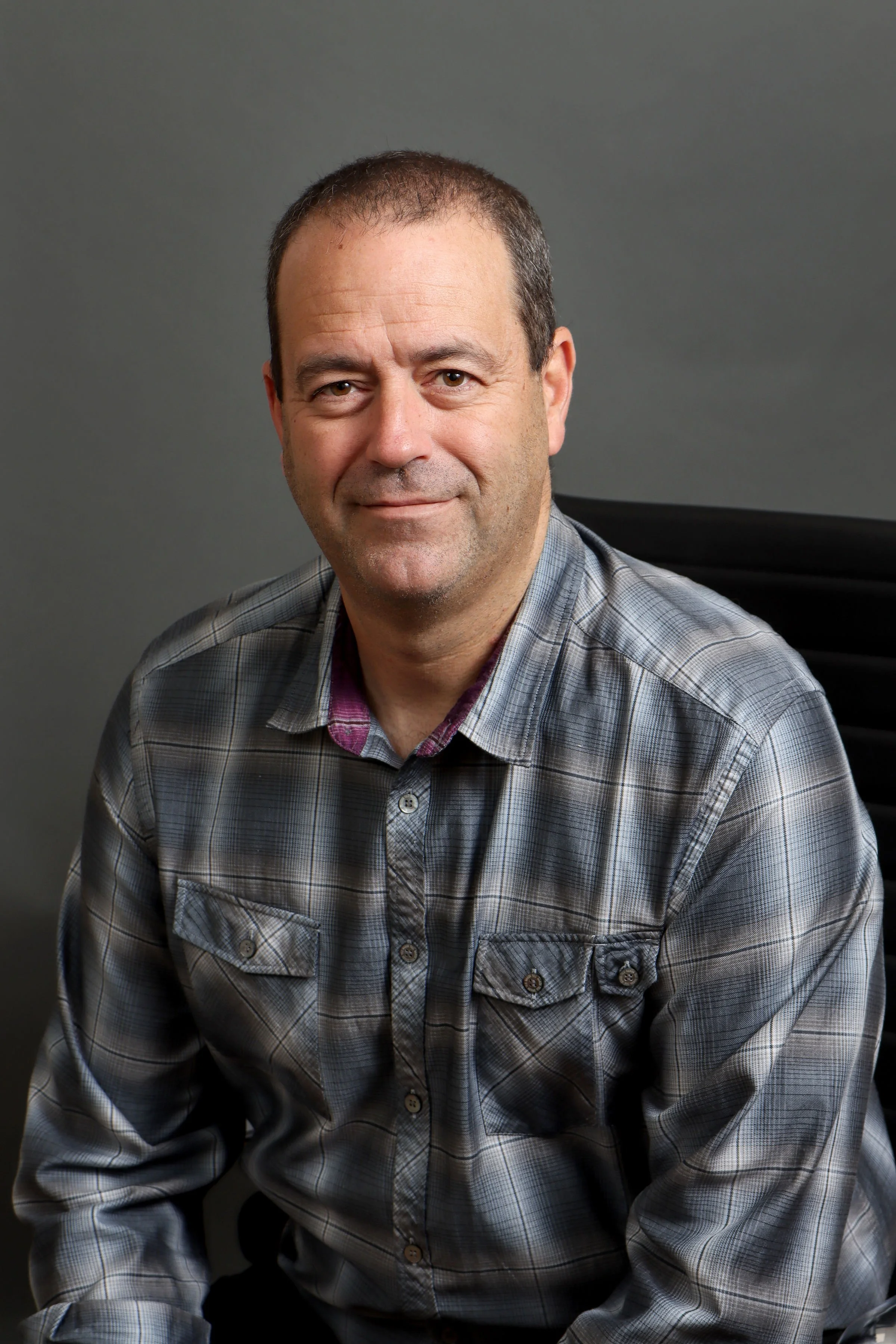 A middle-aged man with short, dark hair and light skin, wearing a gray plaid button-up shirt with a hint of a red and black checkered shirt underneath. He is sitting against a plain, dark gray background and is smiling softly at the camera.