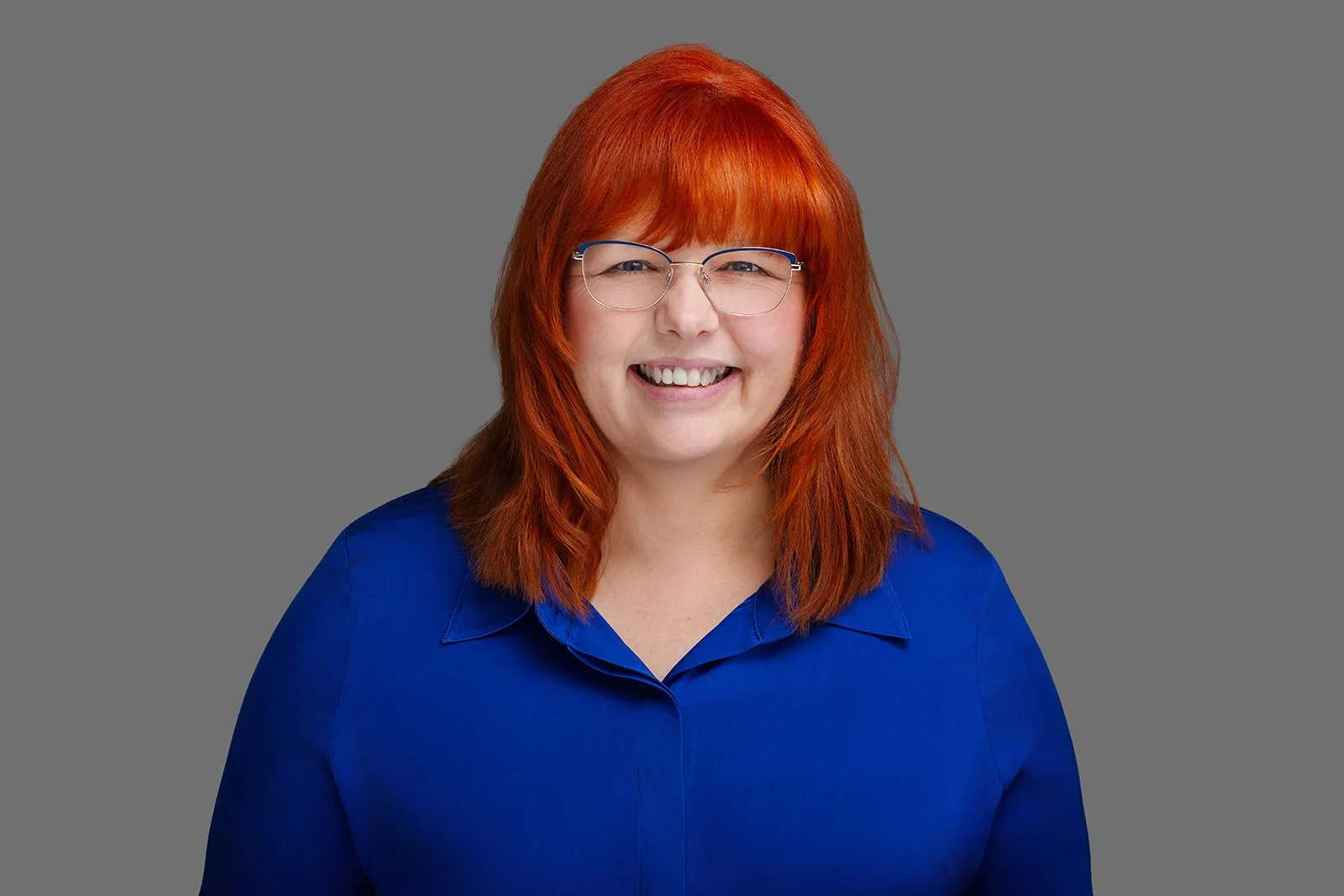 Portrait of a woman with red hair and glasses wearing a blue shirt, smiling against a gray background.