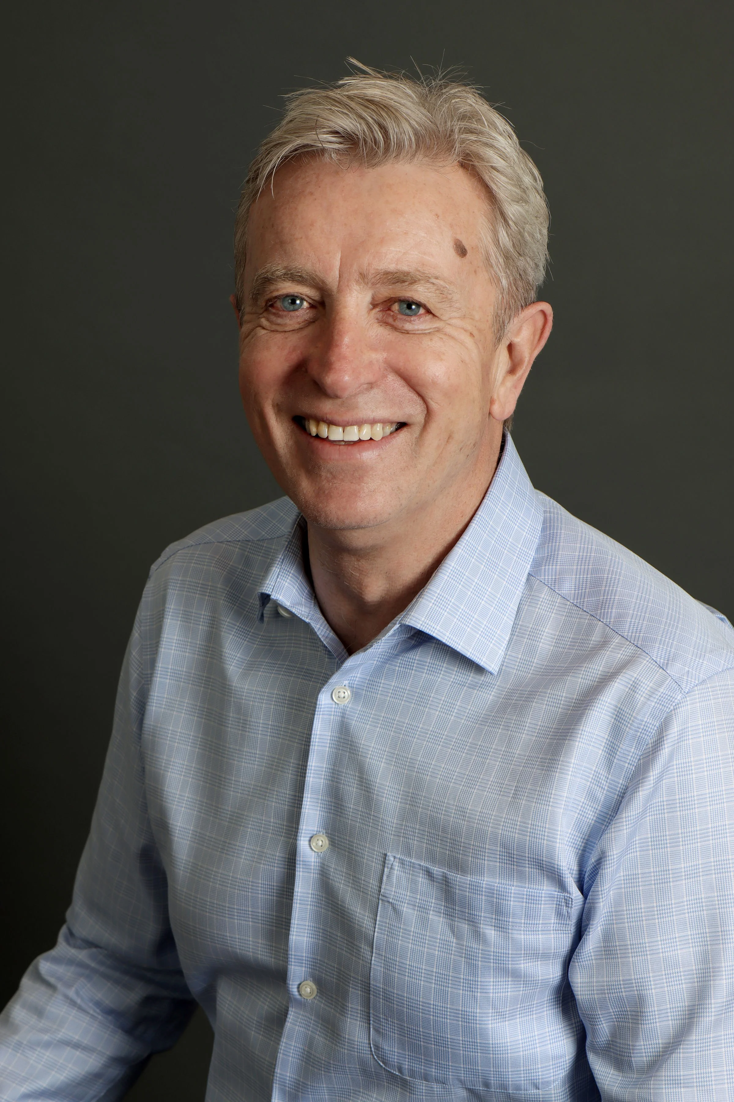 A professional middle-aged man with gray hair, blue eyes, and light skin, smiling, wearing a light blue plaid button-up shirt, against a dark gray background.