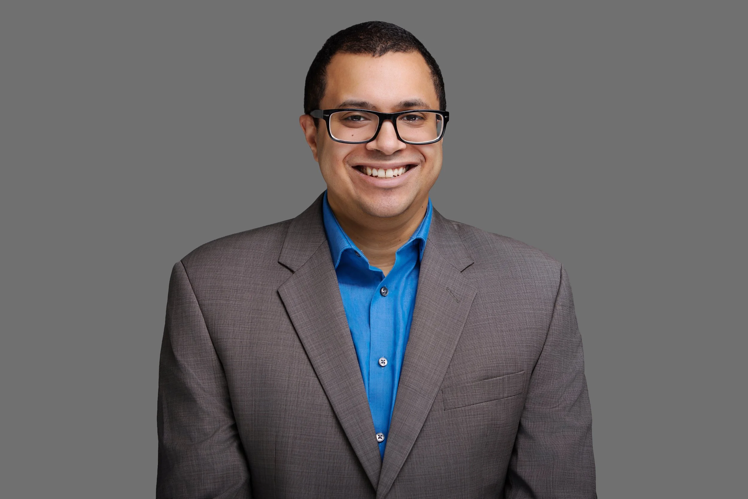 Headshot of a smiling man wearing glasses, a gray blazer, and a blue shirt against a neutral gray background.