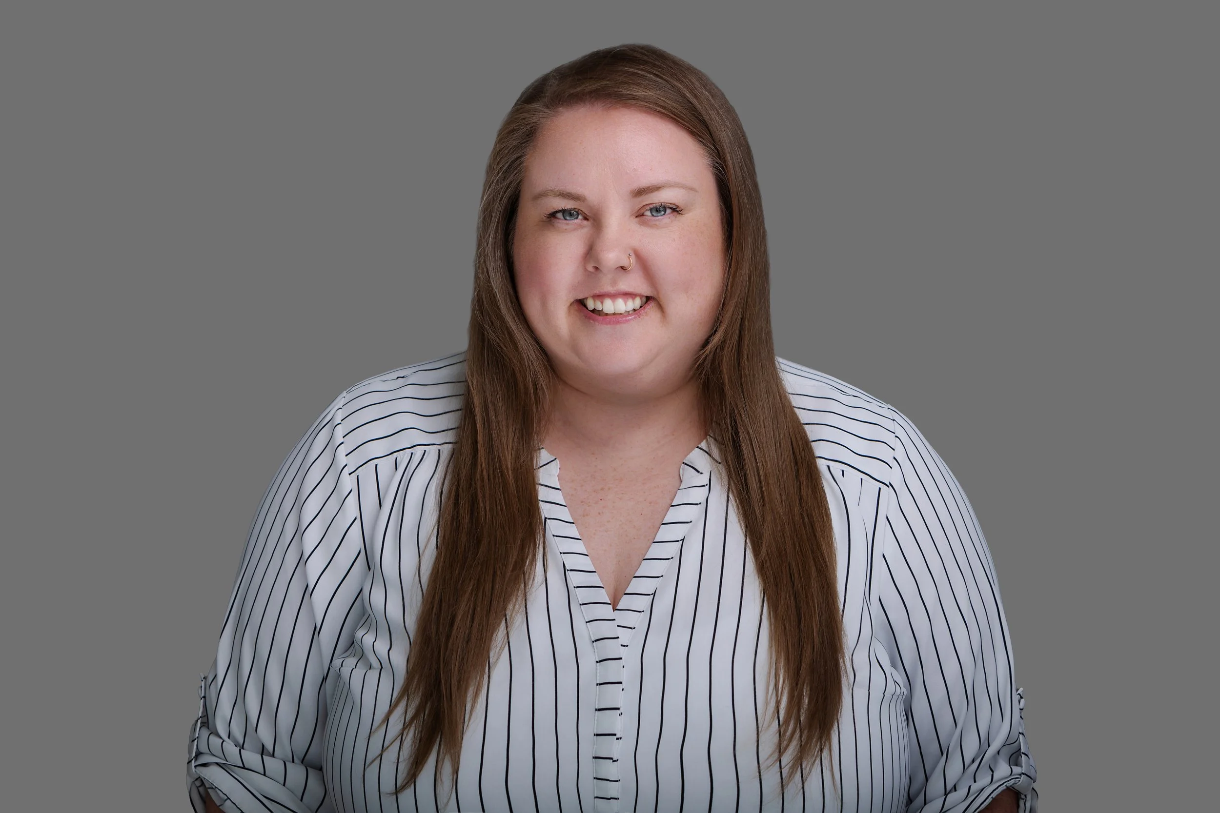 A woman with long brown hair, blue eyes, and a nose piercing smiling at the camera, wearing a white shirt with black vertical pinstripes, against a plain gray background.
