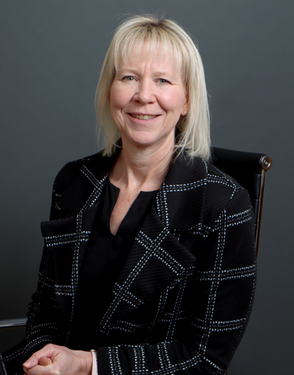 A smiling middle-aged woman with blonde hair, wearing a black top and a black jacket with white checkered stitching, seated on a black chair against a gray background.
