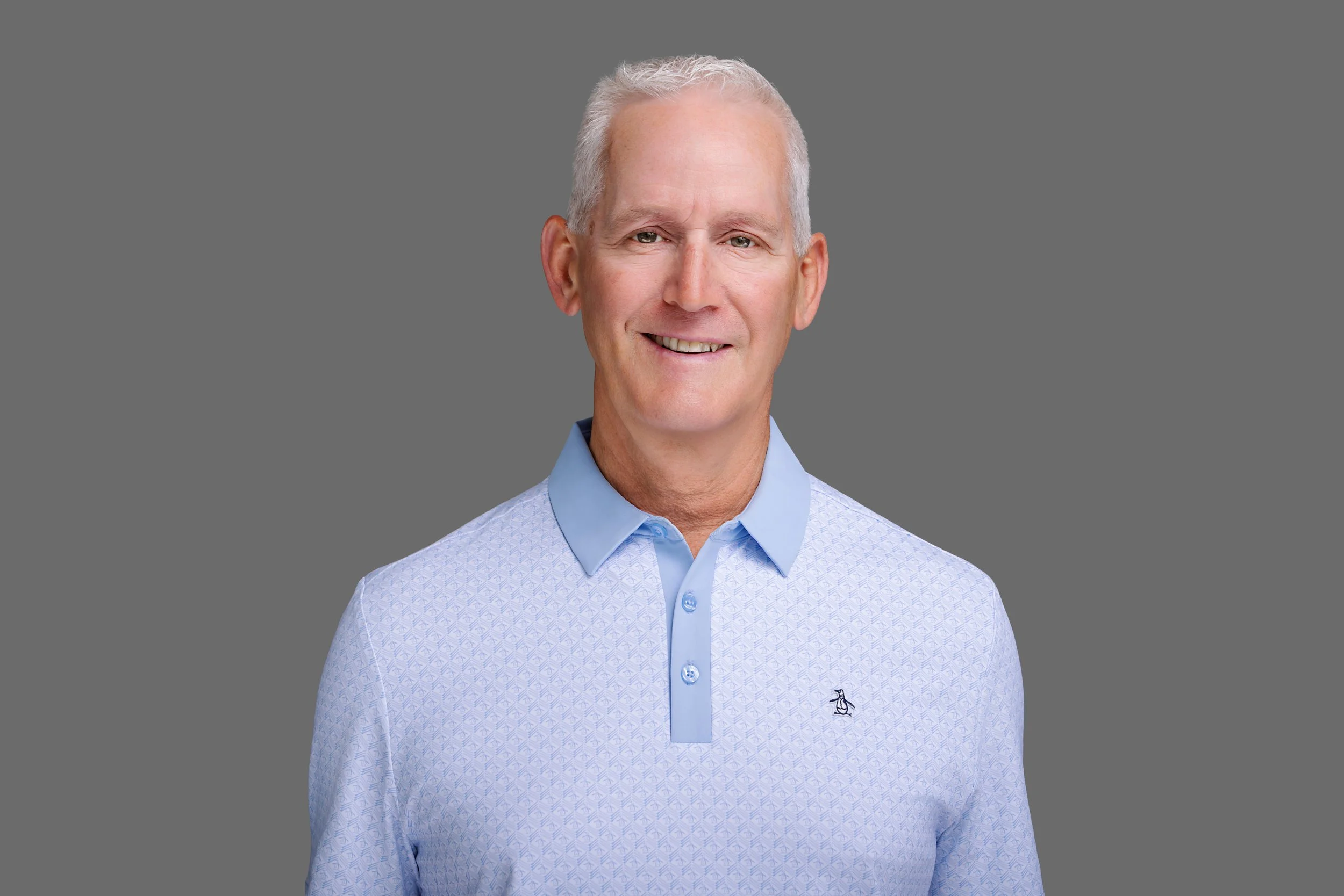 A middle-aged man with short gray hair, wearing a light blue button-up shirt with a small penguin logo on the left chest, stands against a plain gray background, smiling softly.
