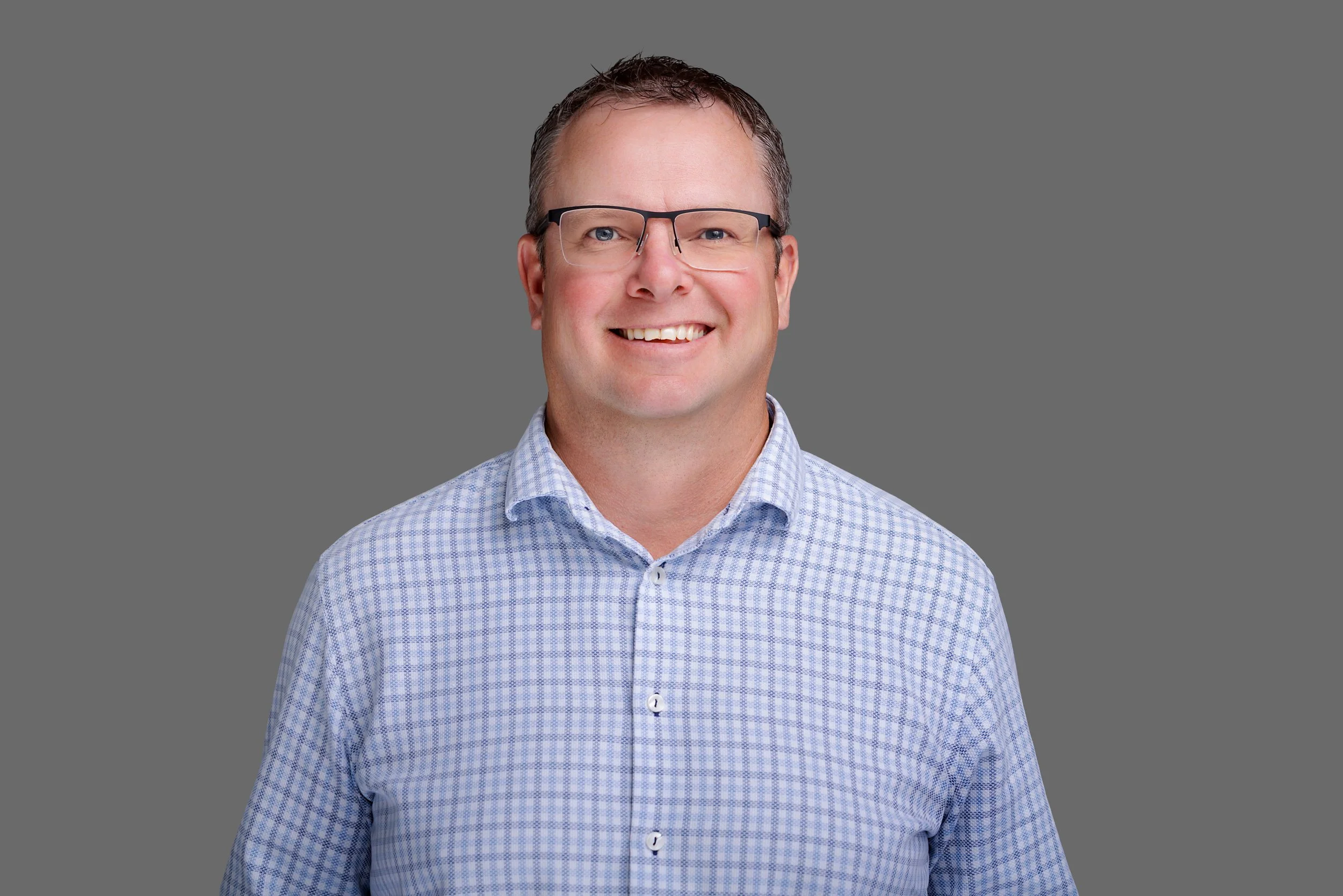 A man smiling, wearing glasses and a light blue checkered shirt, against a plain grey background.
