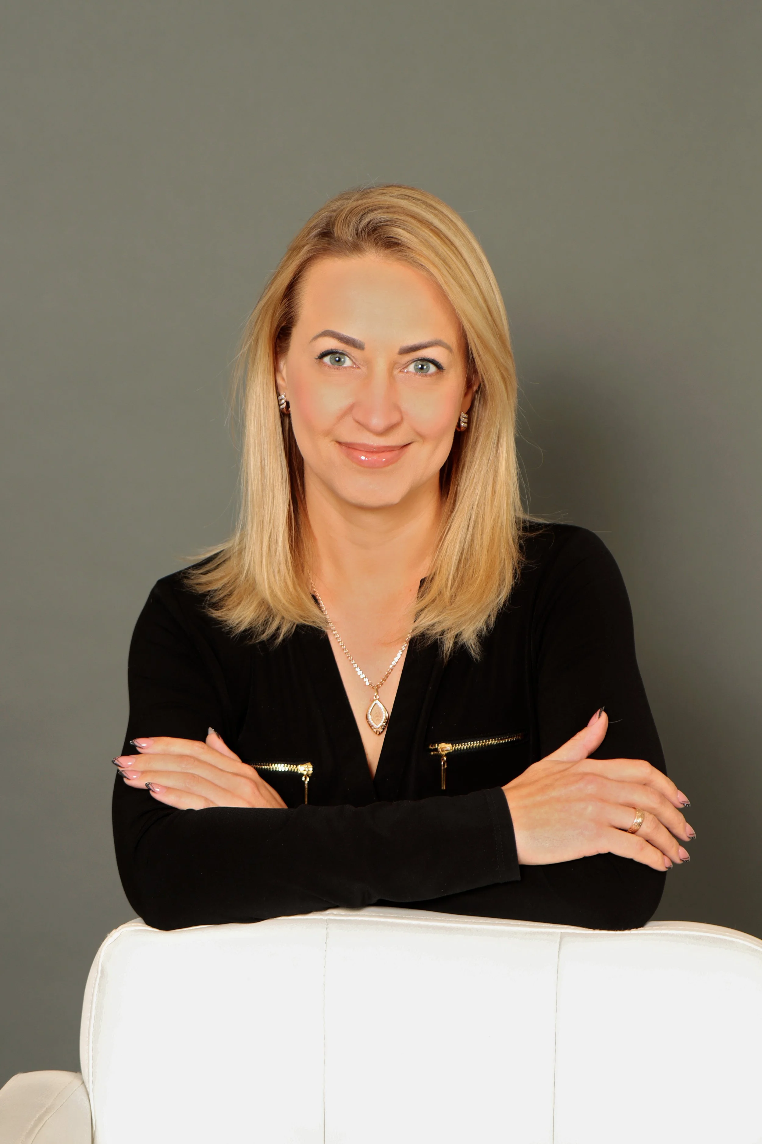 A woman with blonde hair, wearing a black jacket with zippered pockets, silver jewelry, and makeup, seated with her arms crossed, smiling softly against a plain gray background.