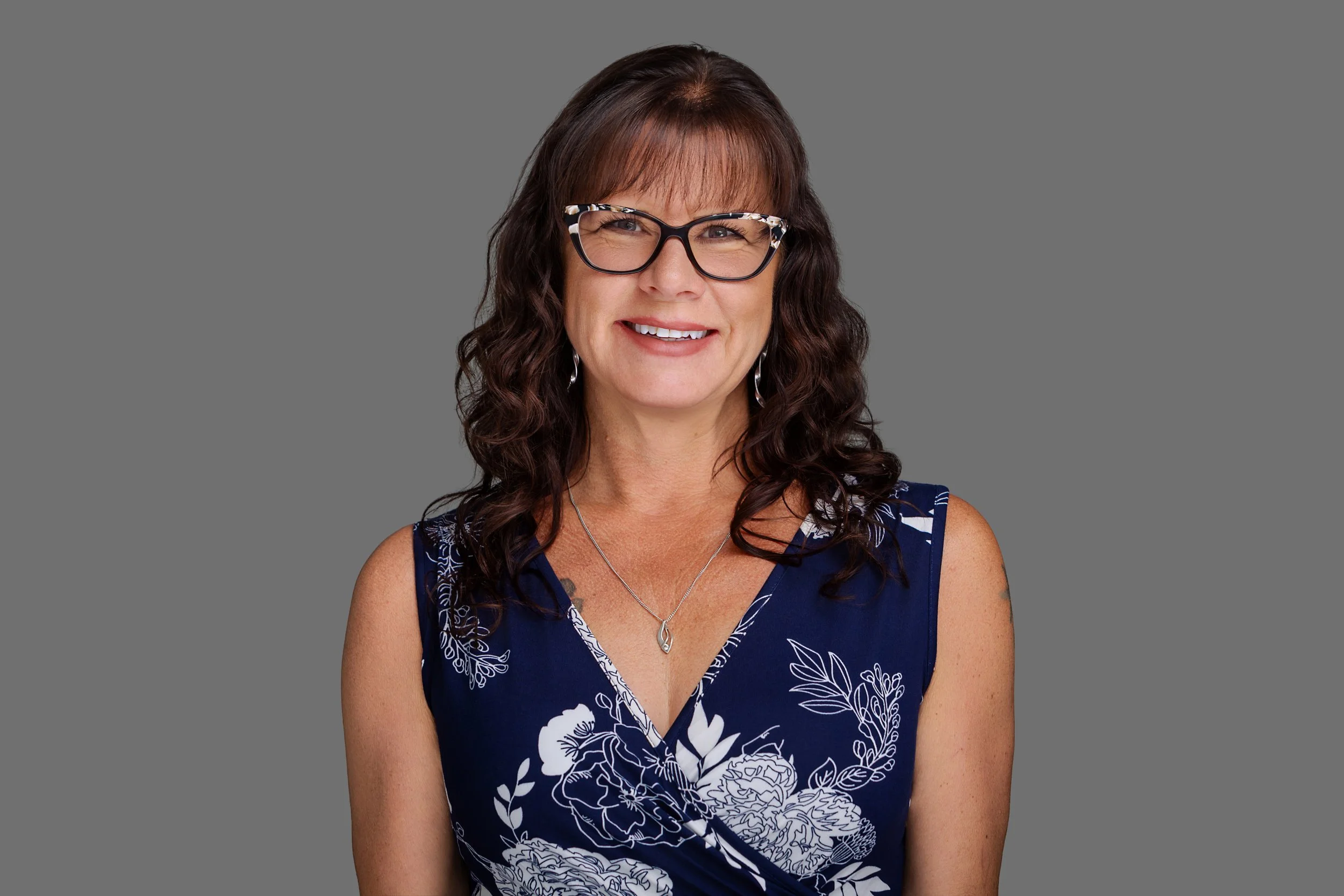 A woman with wavy brown hair, wearing black and white patterned glasses, a blue sleeveless dress with white floral patterns, and a silver necklace, smiling against a plain gray background.